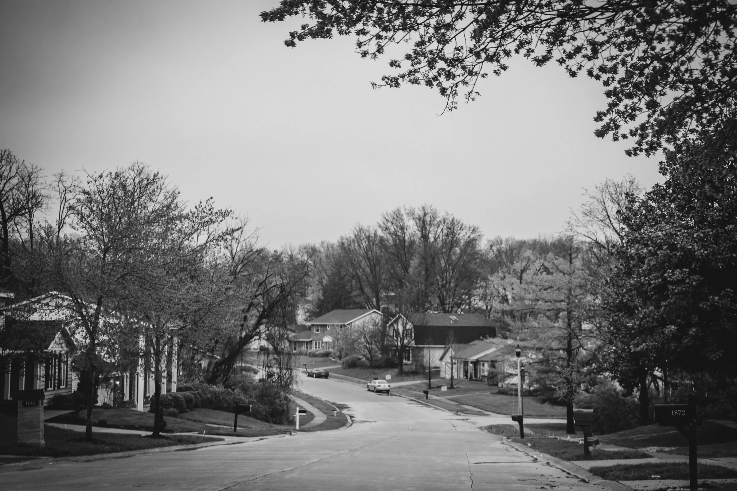 A black and white photo of a neighborhood street with houses lining both sides, leafless trees, and a car driving down the street, viewed from the top of the hill.