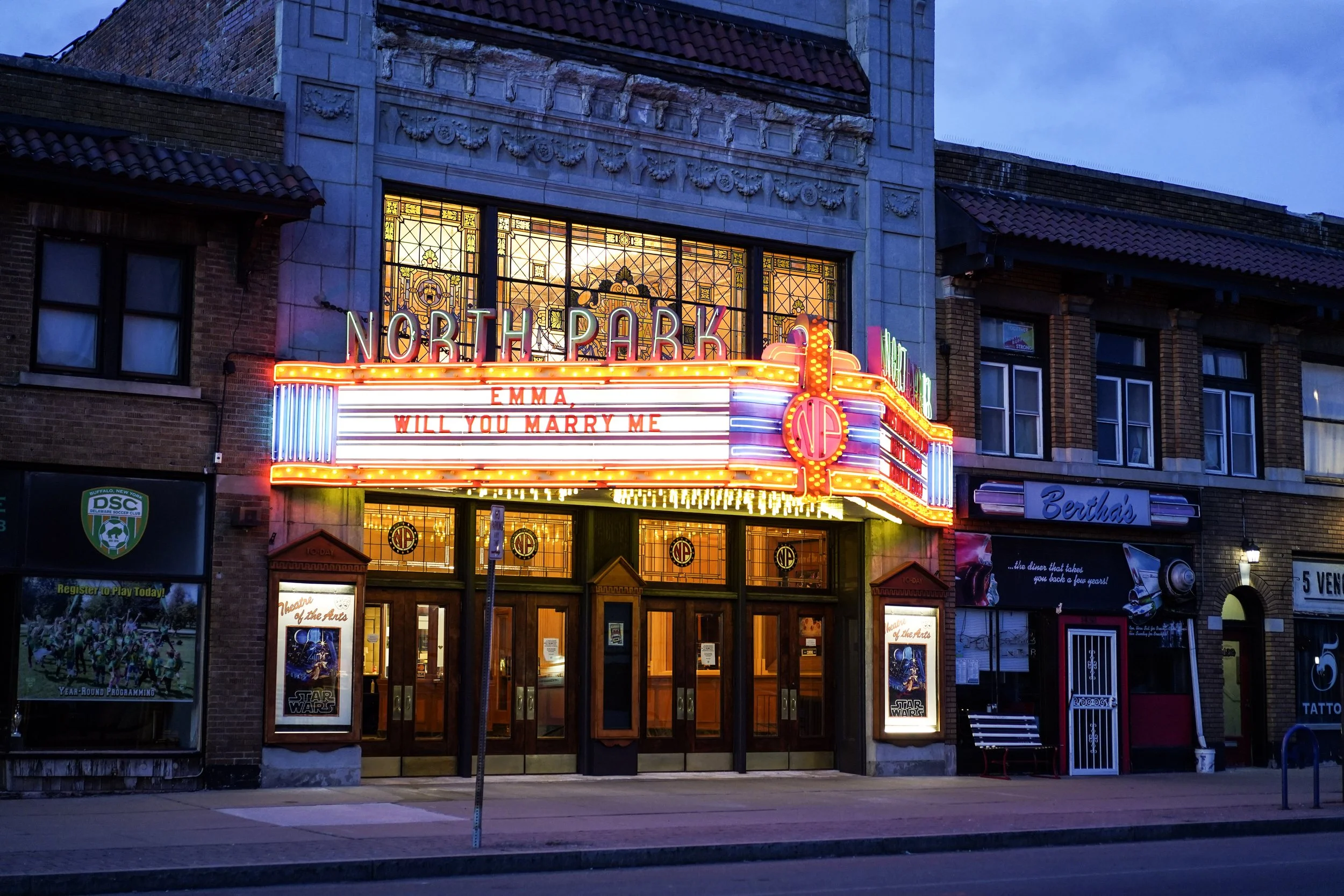 The exterior of the North Park theater with a neon sign, marquee, and posters, during twilight. The marquee displays the words 'Emma' and 'Will You Marry Me'.