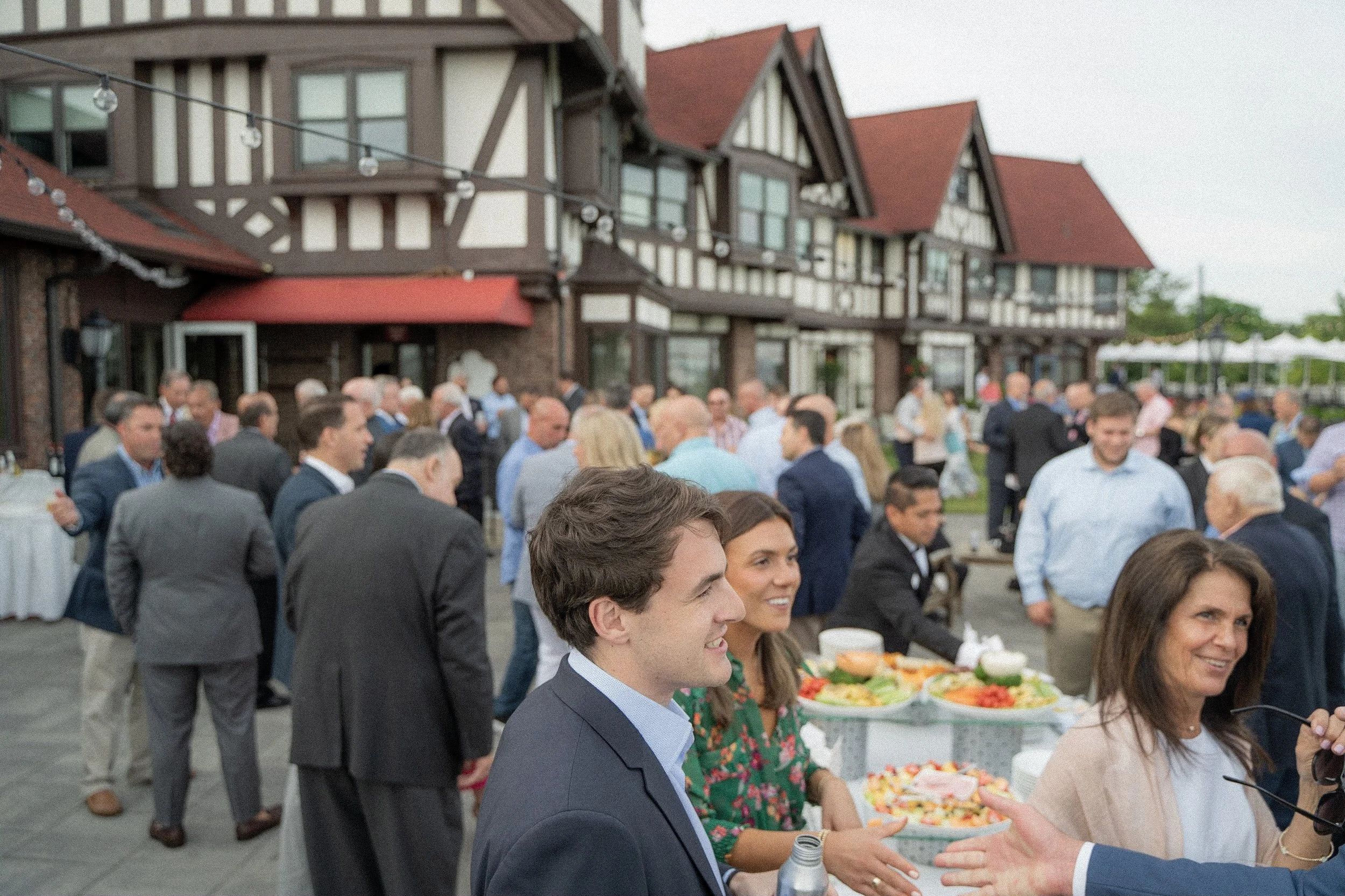 People gathered outdoors at a social event or party with a large crowd, some serving food on tables, in front of a large Tudor-style house with string lights hanging overhead.