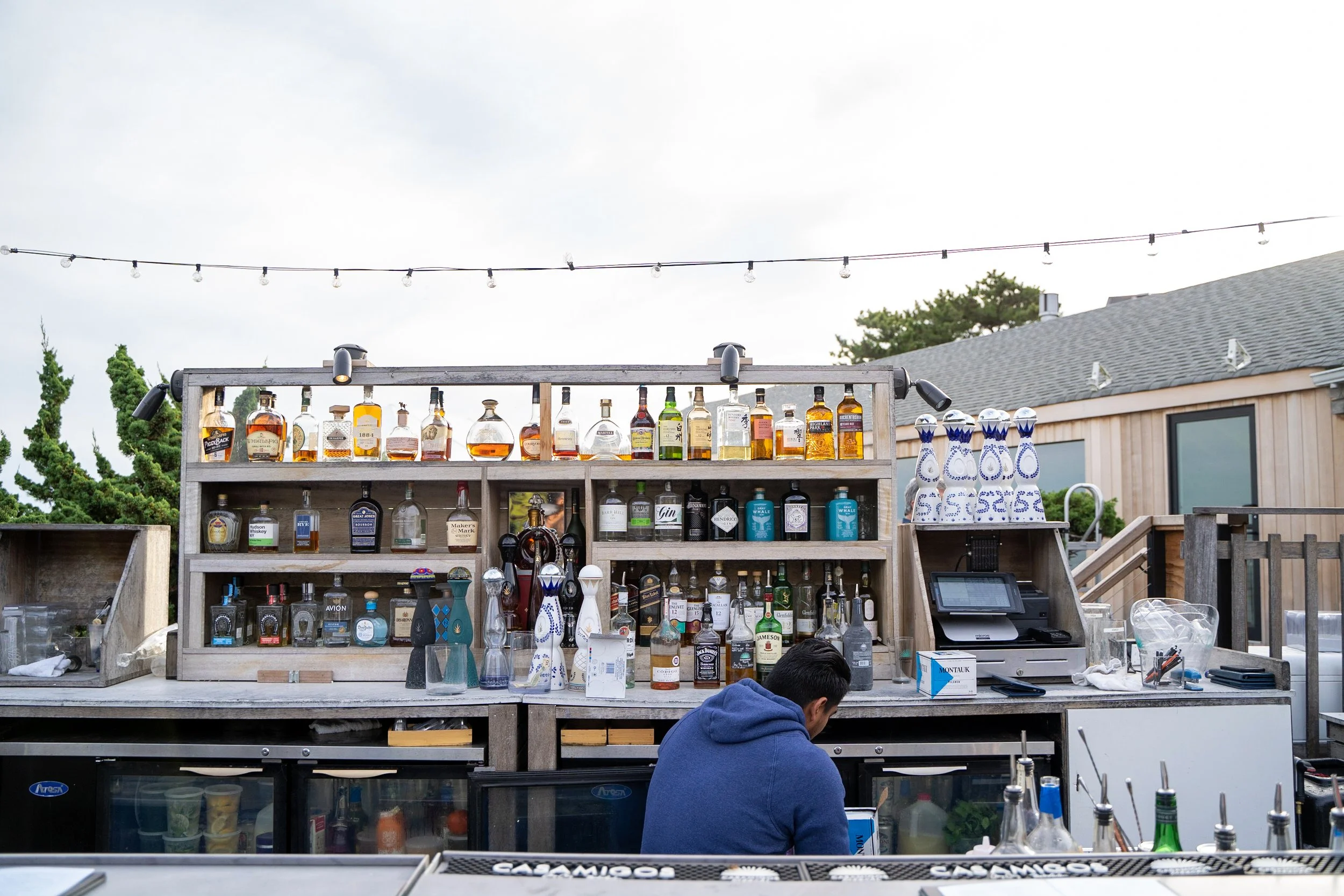 Outdoor bar setup with liquor bottles, bar accessories, and a person in a blue hoodie working behind the counter. String lights are overhead and a house with wooden siding is in the background.