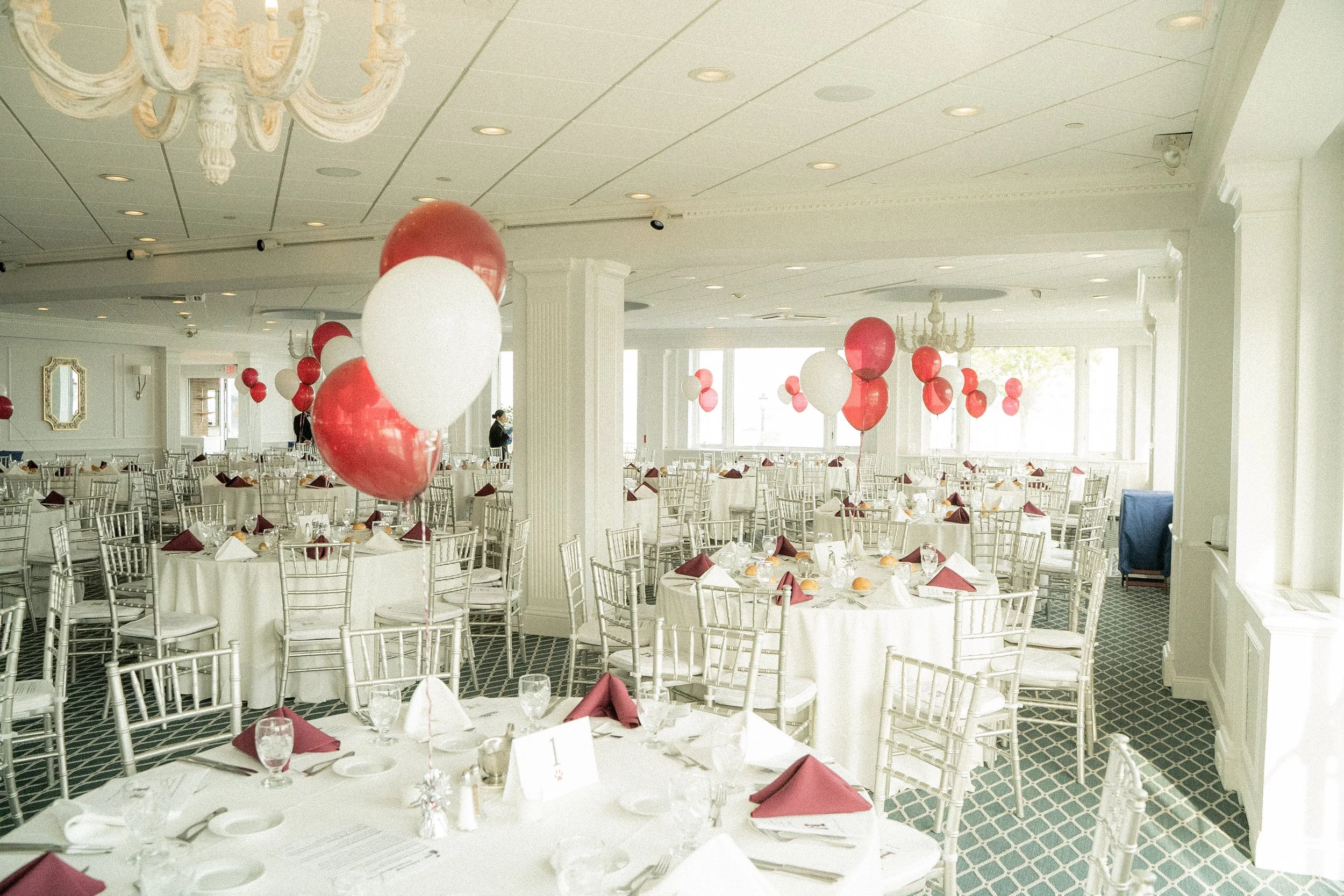 A banquet hall decorated with red and white balloons, set for a formal event with round tables covered in white tablecloths, red napkins, glassware, and plates, with chandeliers hanging from the ceiling.