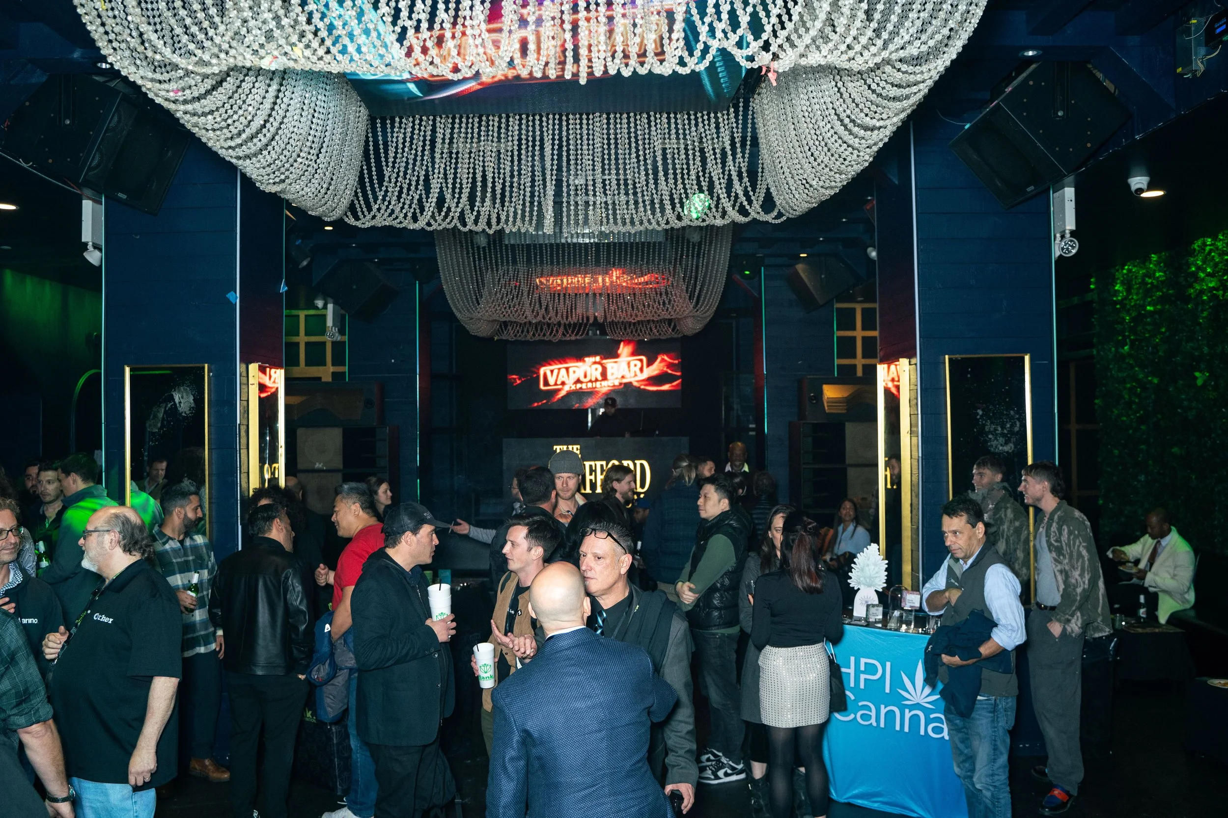 Crowd of people attending a vape bar event inside a modern venue with decorative ceiling lights and a digital sign reading 'Vapor Bar' in the background.