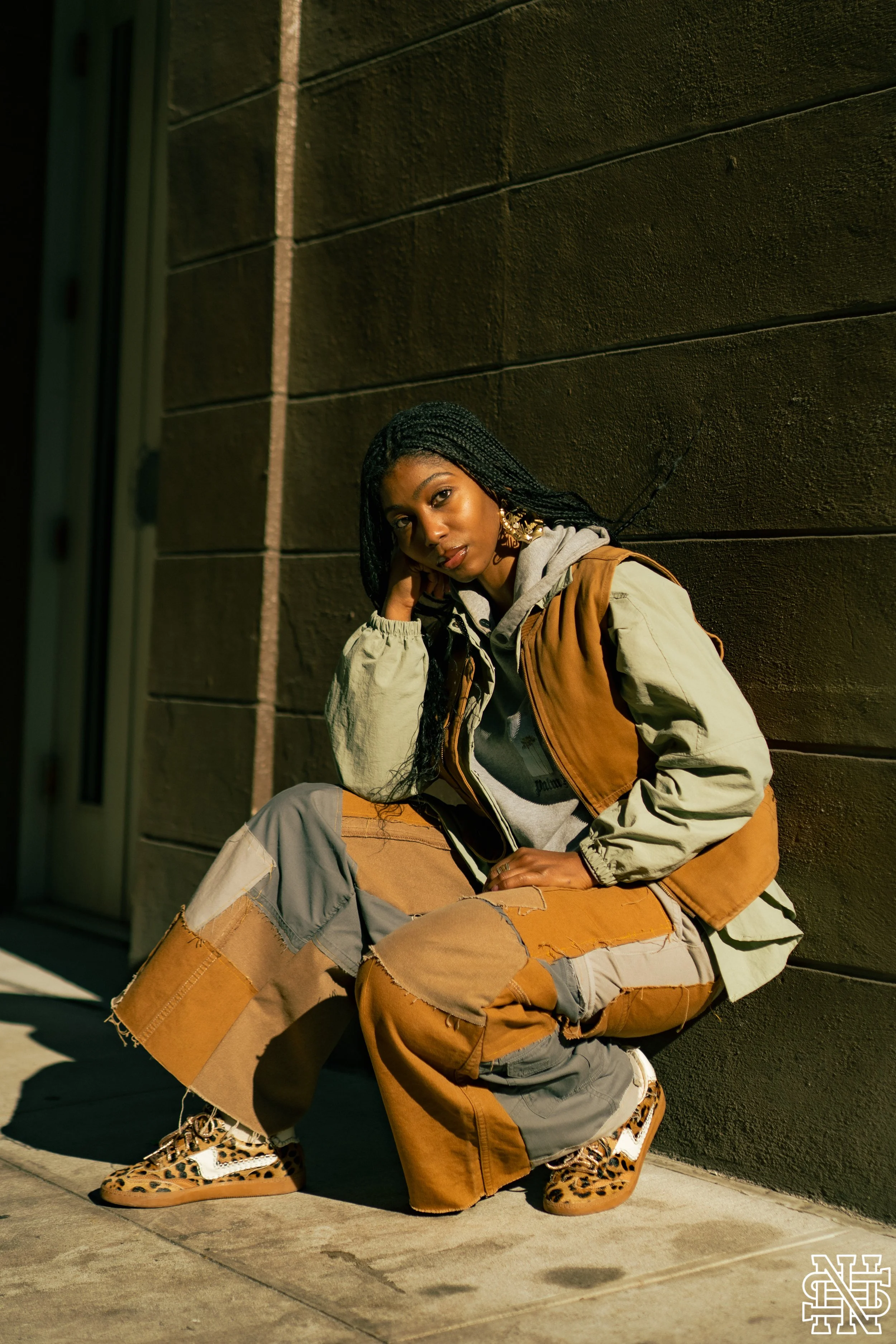 A young woman with long braided hair poses against a dark wall, crouching on the sidewalk with one hand on her face. She is wearing a gray hoodie, a brown and beige color-blocked jacket, baggy patchwork pants, and leopard print sneakers. The lighting
