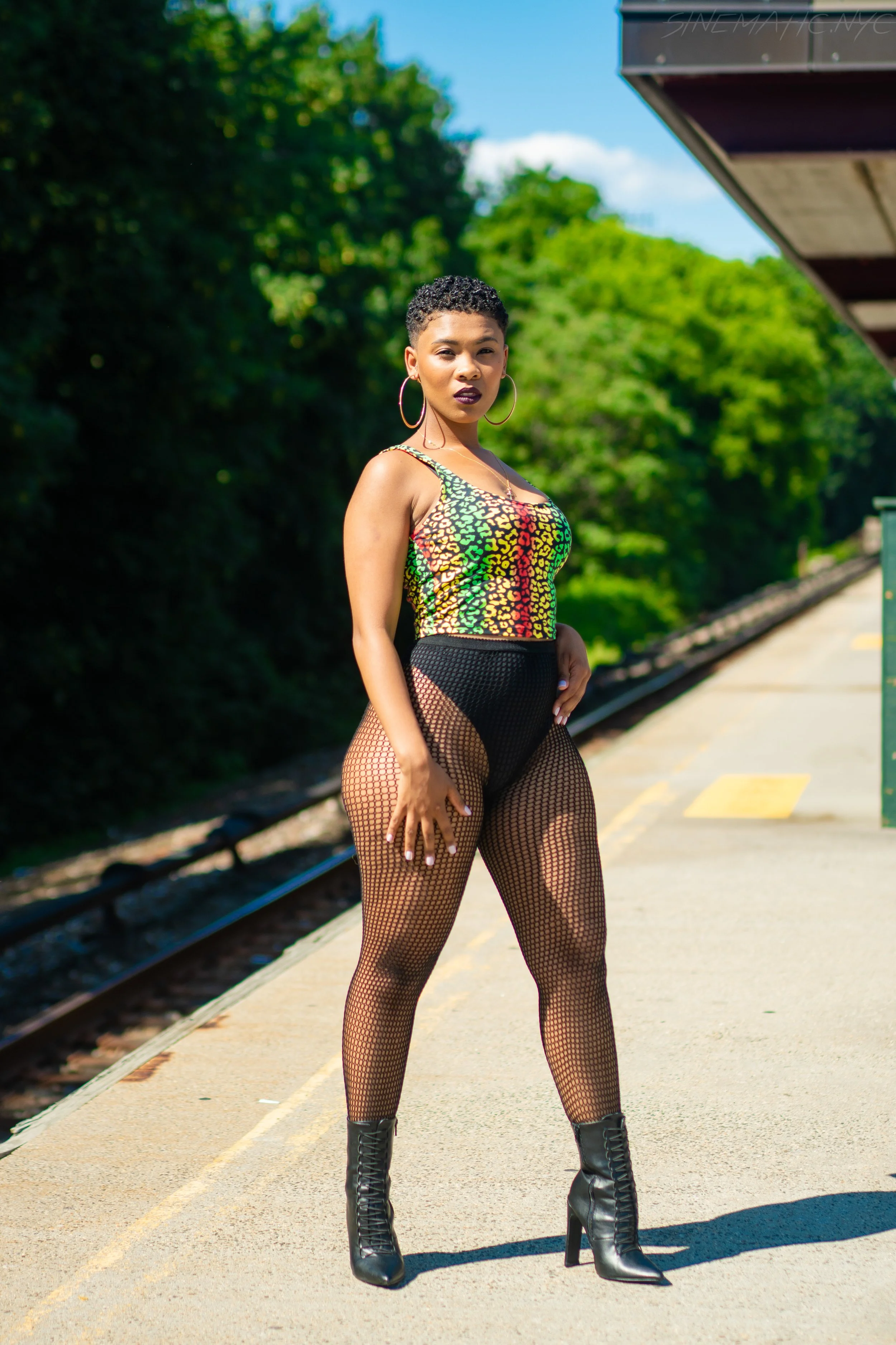 Woman standing on train platform wearing colorful animal print top, black fishnet tights, and black high-heeled boots.