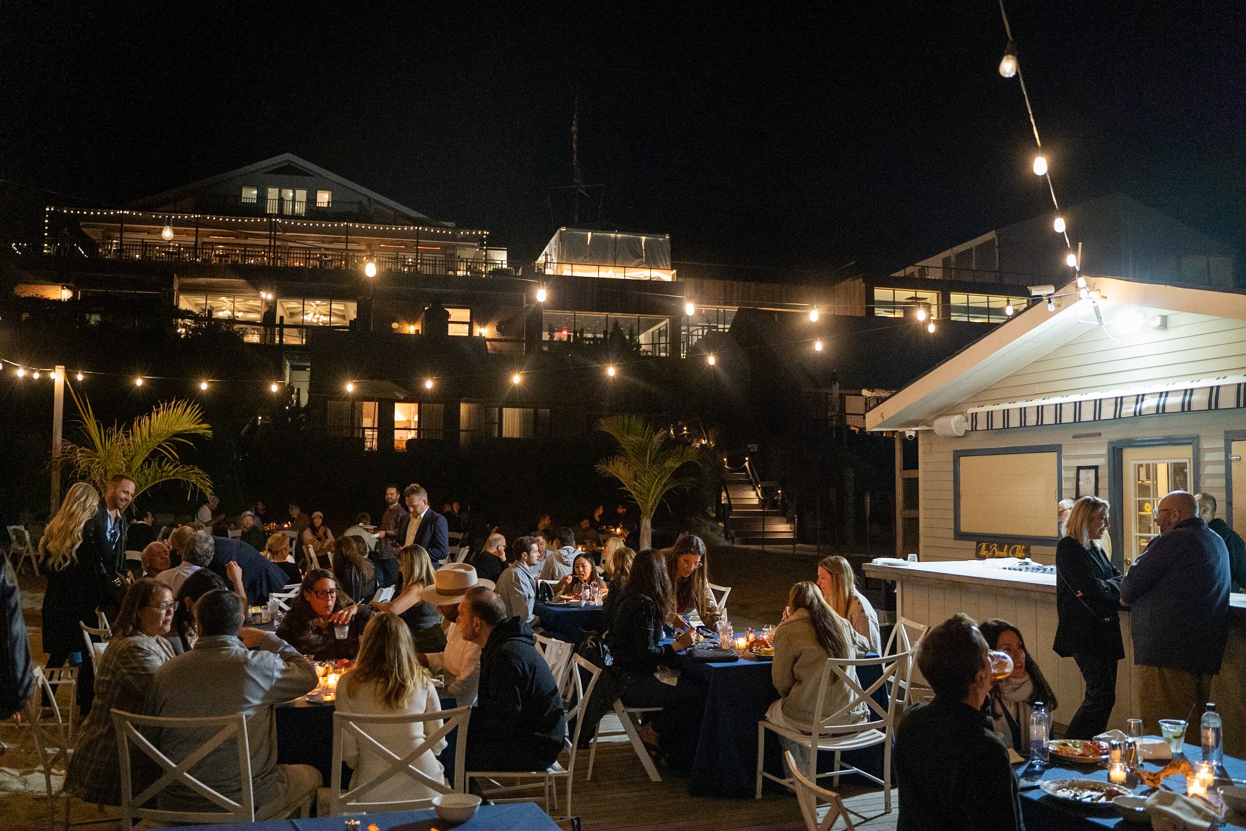 Outdoor nighttime scene of a social gathering with people dining at tables, string lights above, and houses or buildings lit up in the background.