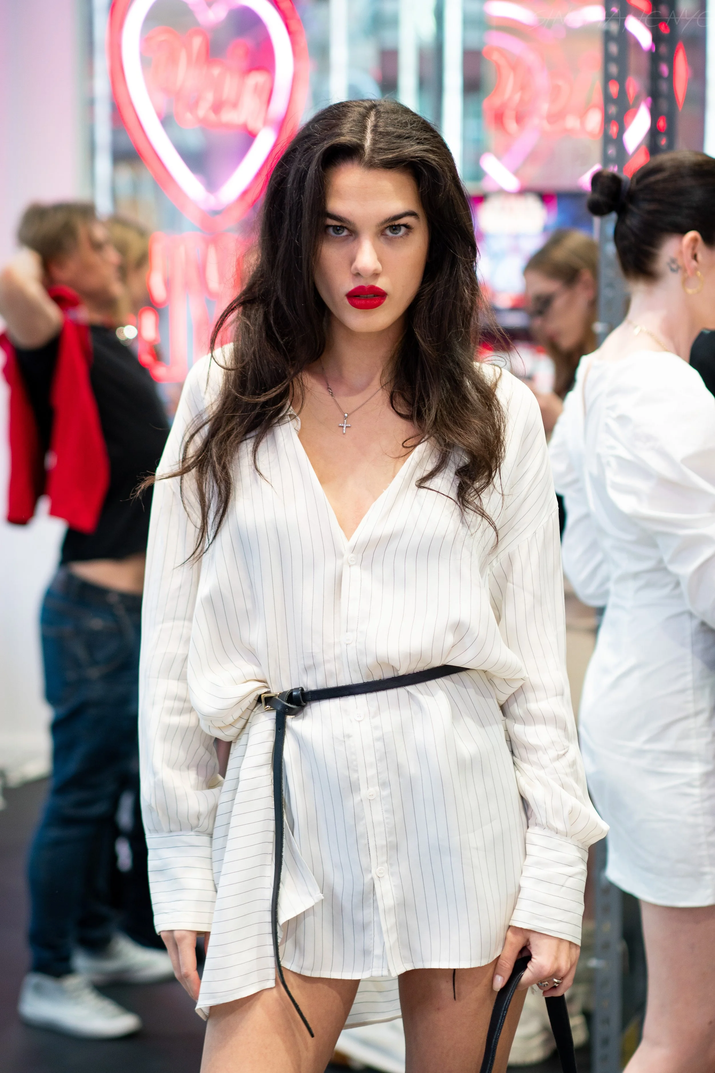 A young woman with long dark hair and red lipstick stands indoors with several people in the background. She wears a white, long-sleeve, button-up shirt with thin vertical stripes, tied at the waist with a black belt, revealing her legs. Neon signs a