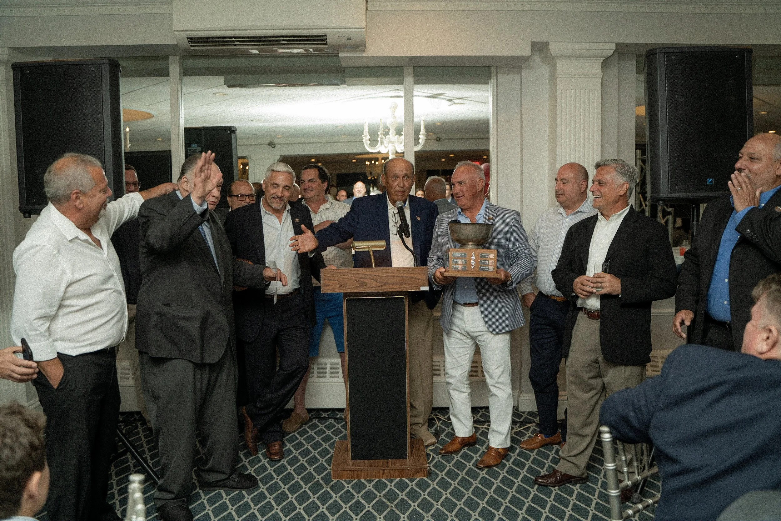 Group of men in formal attire at a celebration, with one man speaking at a podium and others smiling and engaging, some holding a trophy and a glass in a banquet hall.