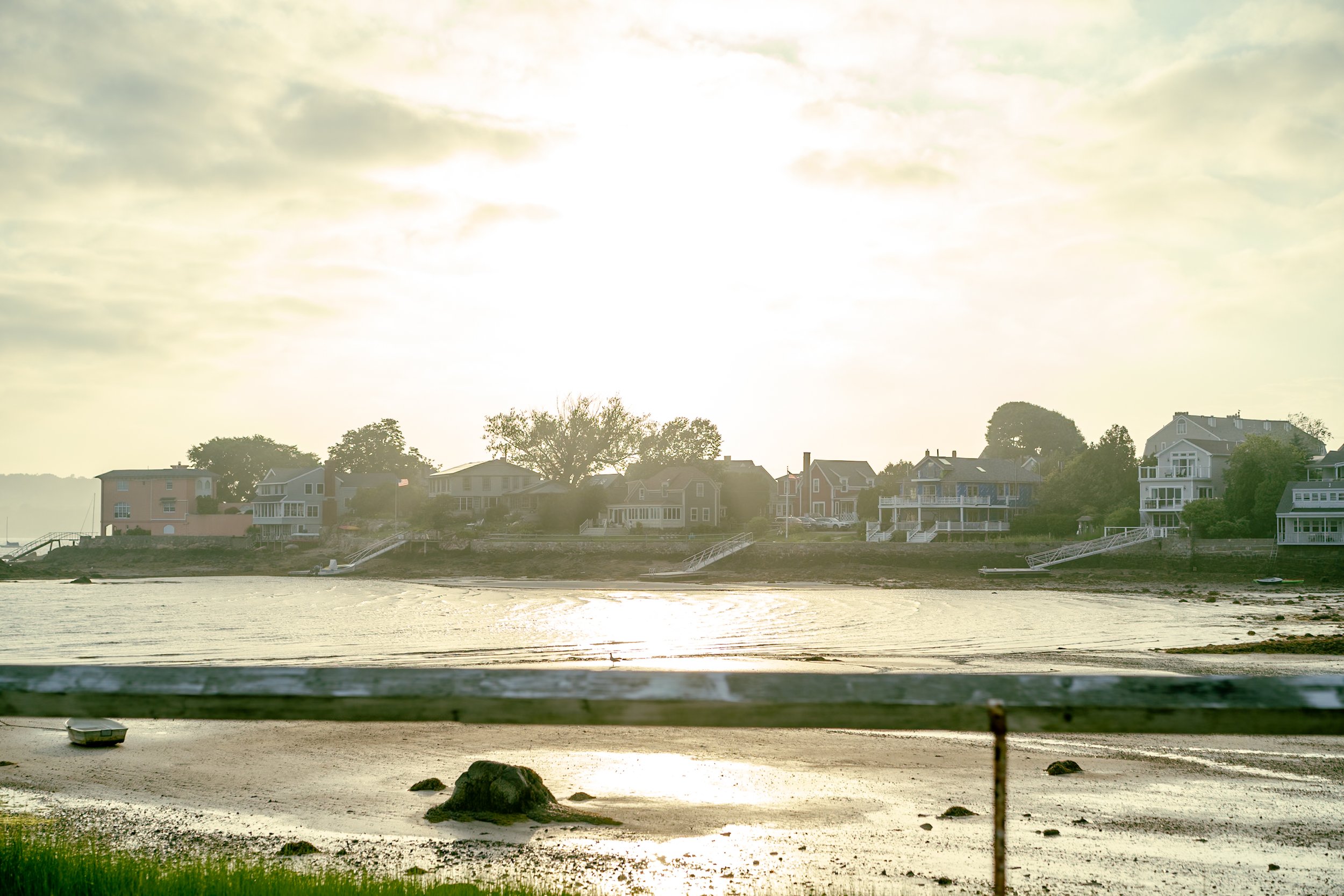 Houses on a shoreline at sunset, with a calm body of water in front and a railing in the foreground.