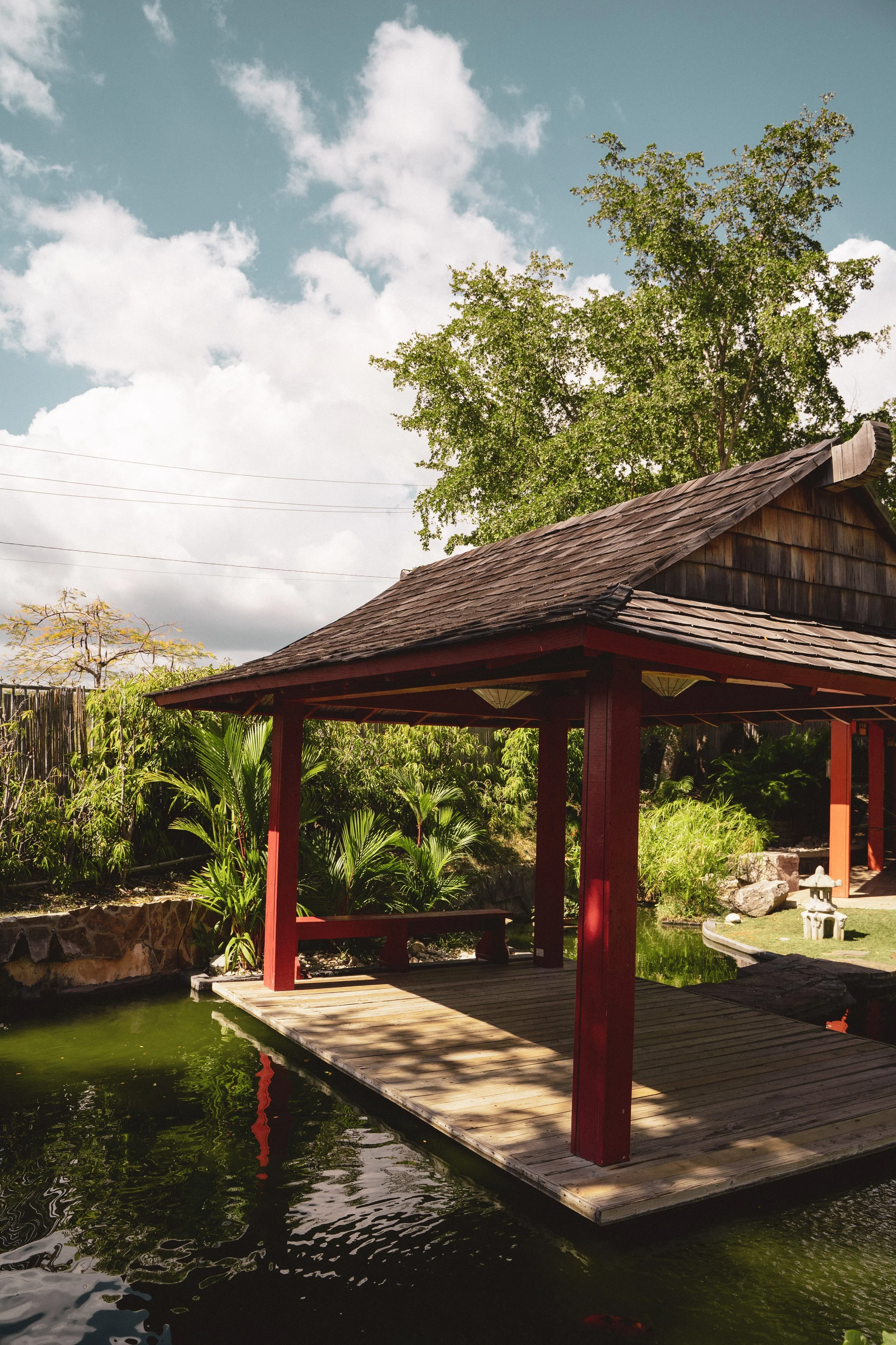 A traditional Asian-style pavilion with a wooden deck built over a pond, surrounded by lush greenery and trees under a partly cloudy sky.