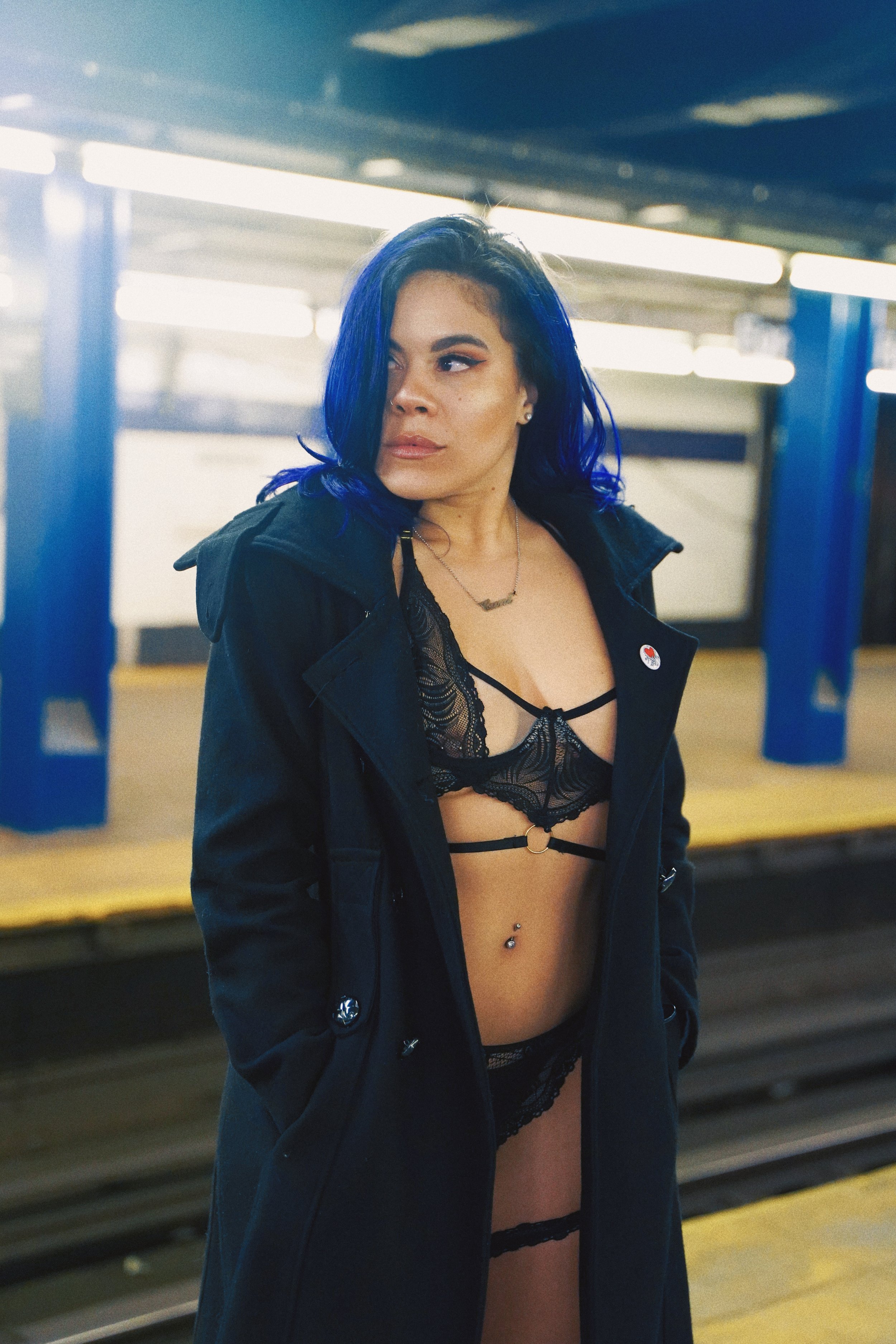A woman with blue hair standing on a train platform, dressed in black lingerie and a long black coat.