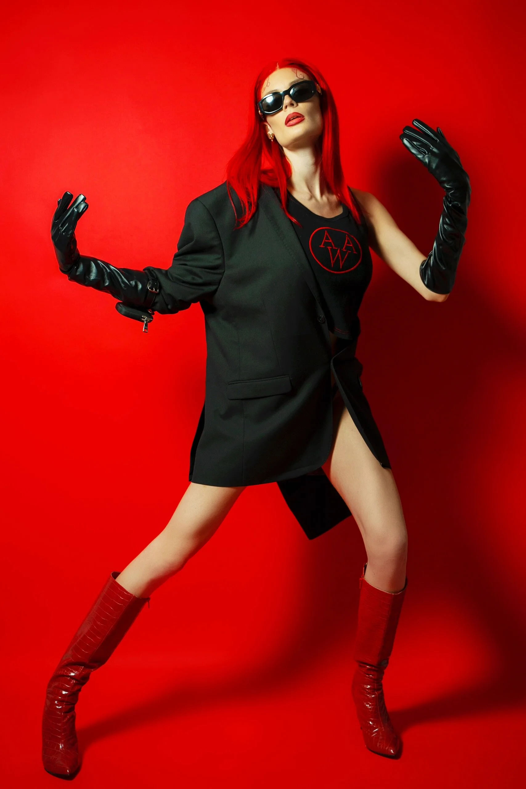 A woman with red hair, sunglasses, and red lipstick posing against a red background. She is dressed in a black blazer and boots, with long black gloves. She is striking a confident pose with her arms raised and legs apart.