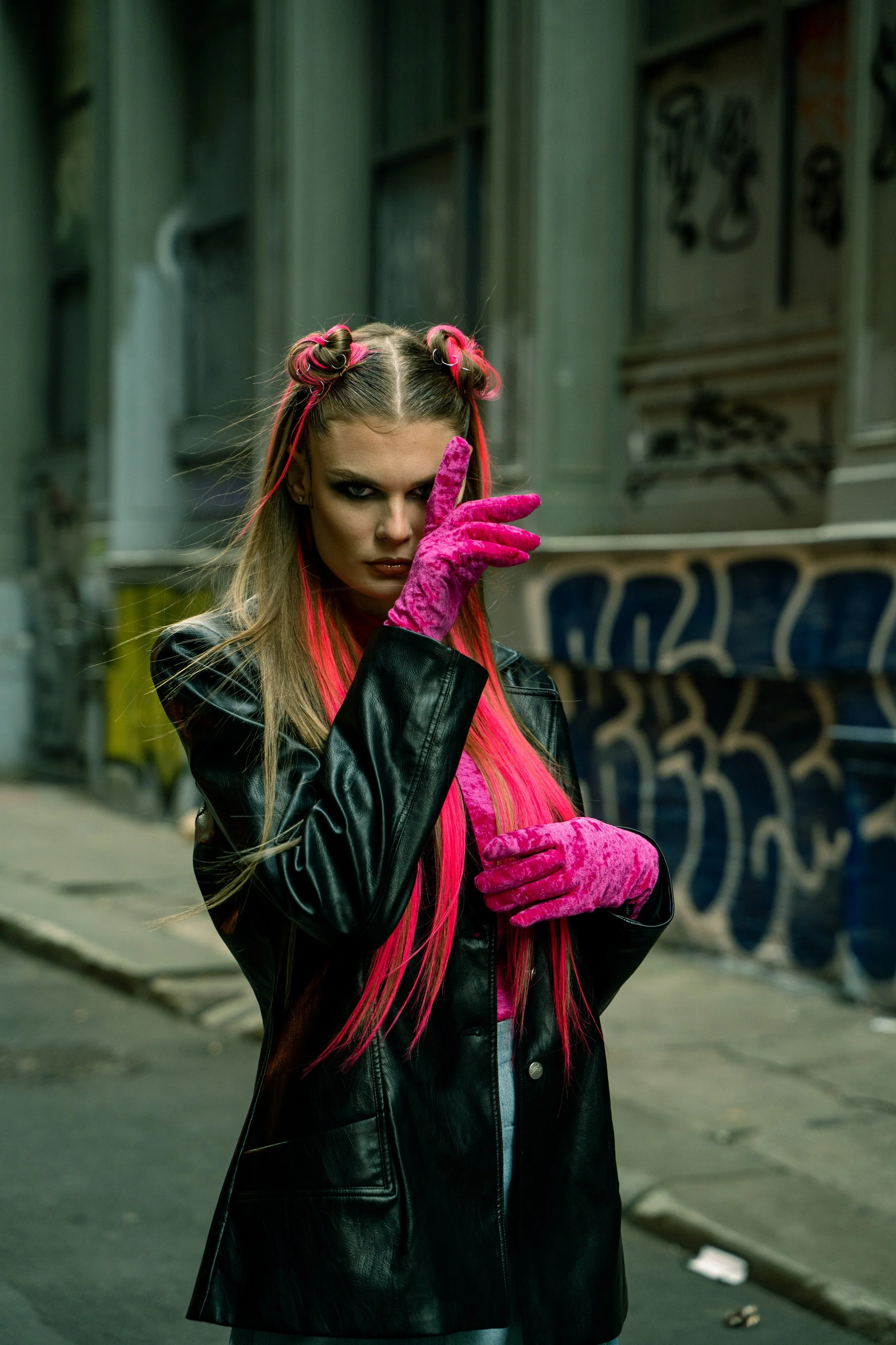 A young woman with long, styled hair and bold makeup poses on an urban street, wearing a black leather jacket and bright pink velvet gloves, with one hand partially covering her face.