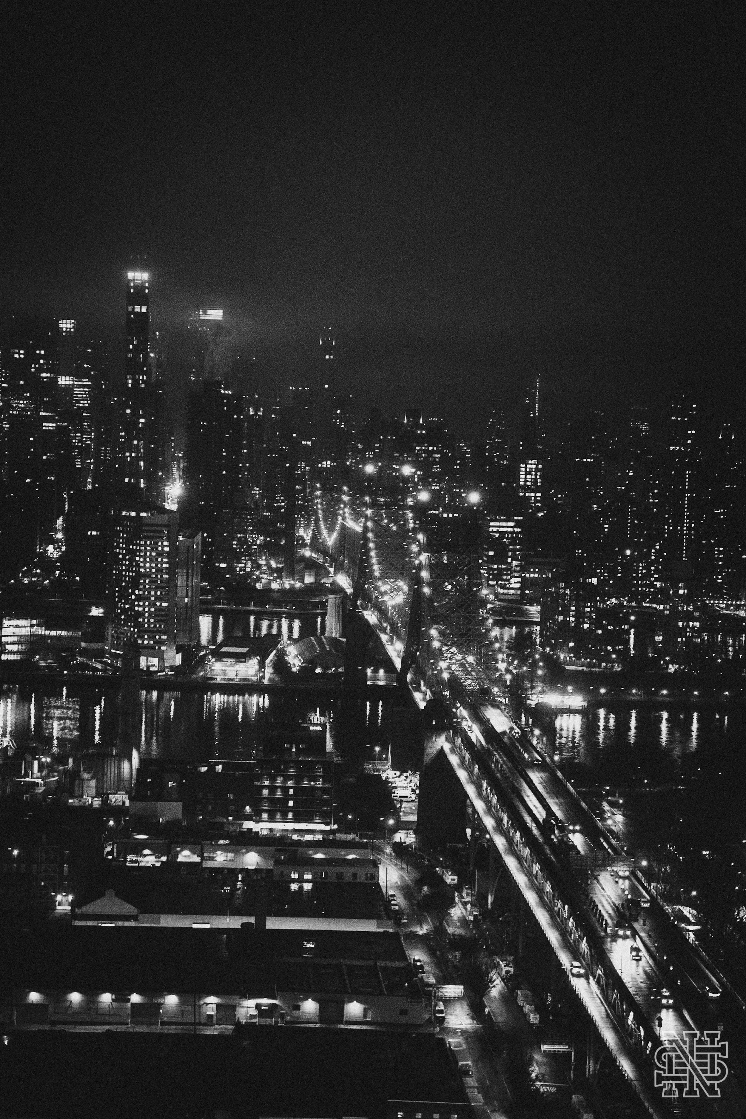 Nighttime cityscape with illuminated skyscrapers, a bridge over water, and a grid of streets with streetlights.