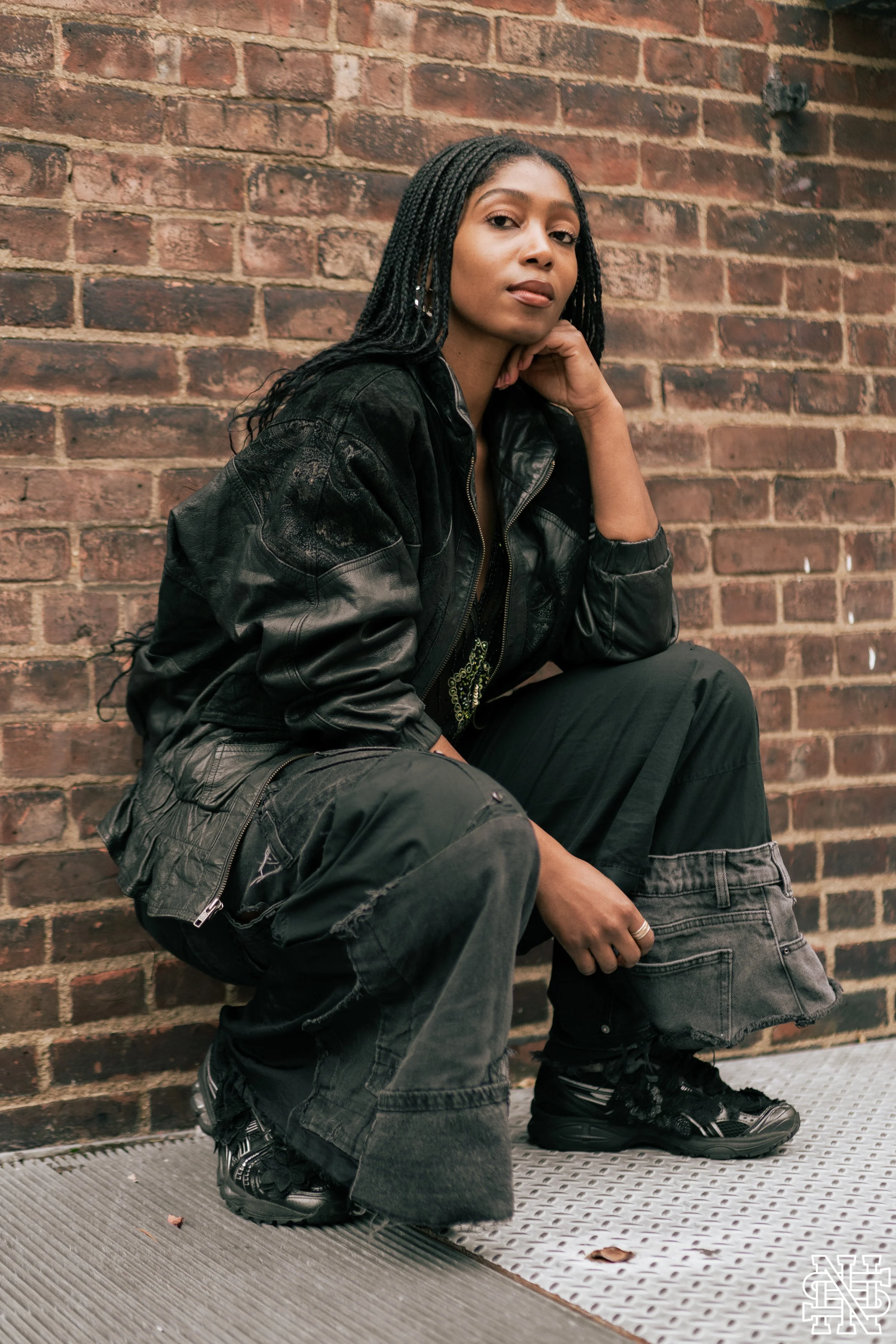 A young woman with box braids wearing a black leather jacket, black pants, and sneakers, sitting against a brick wall, looking at the camera.