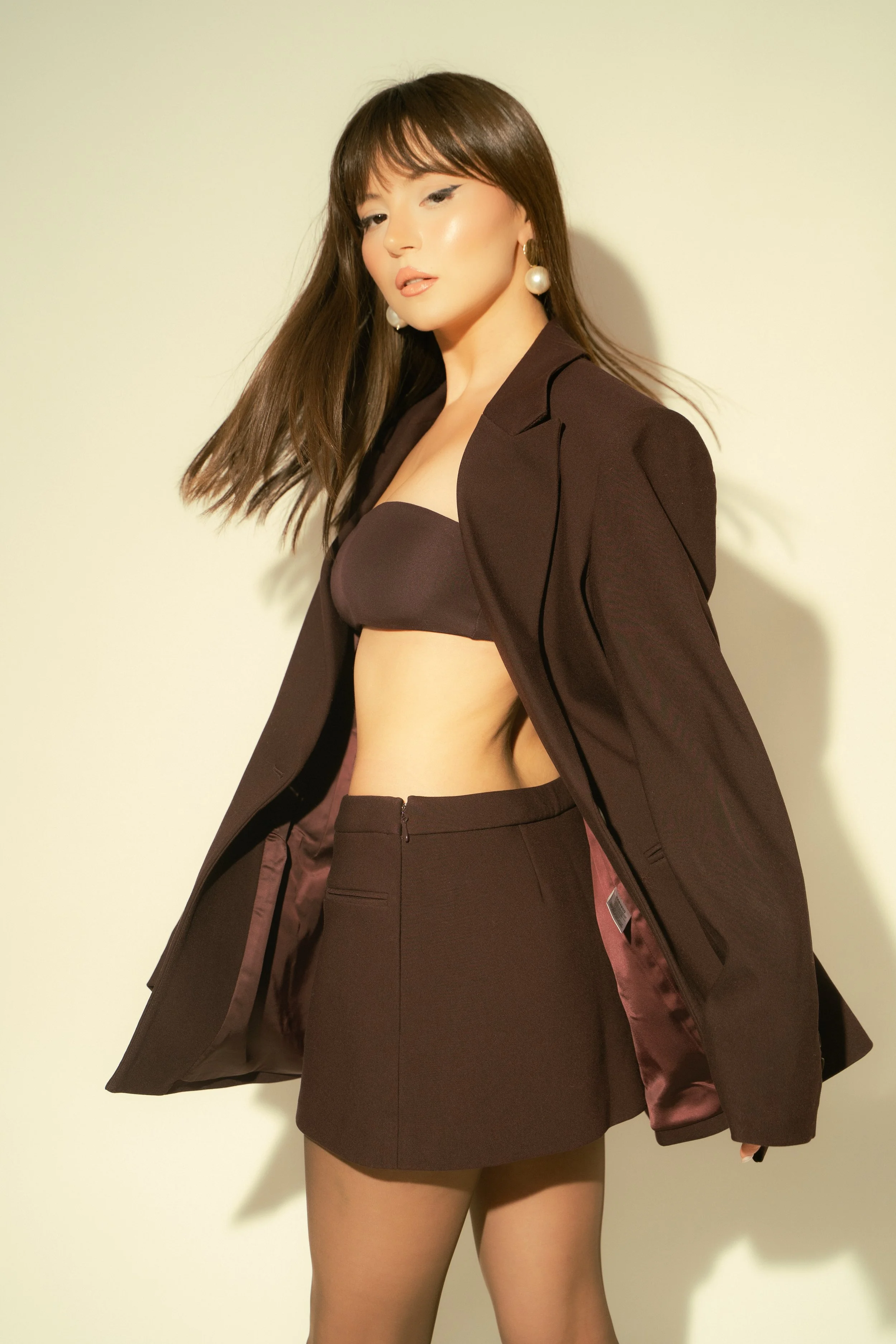 Woman wearing a dark blazer, bandeau top, and skirt, posing against a plain background.