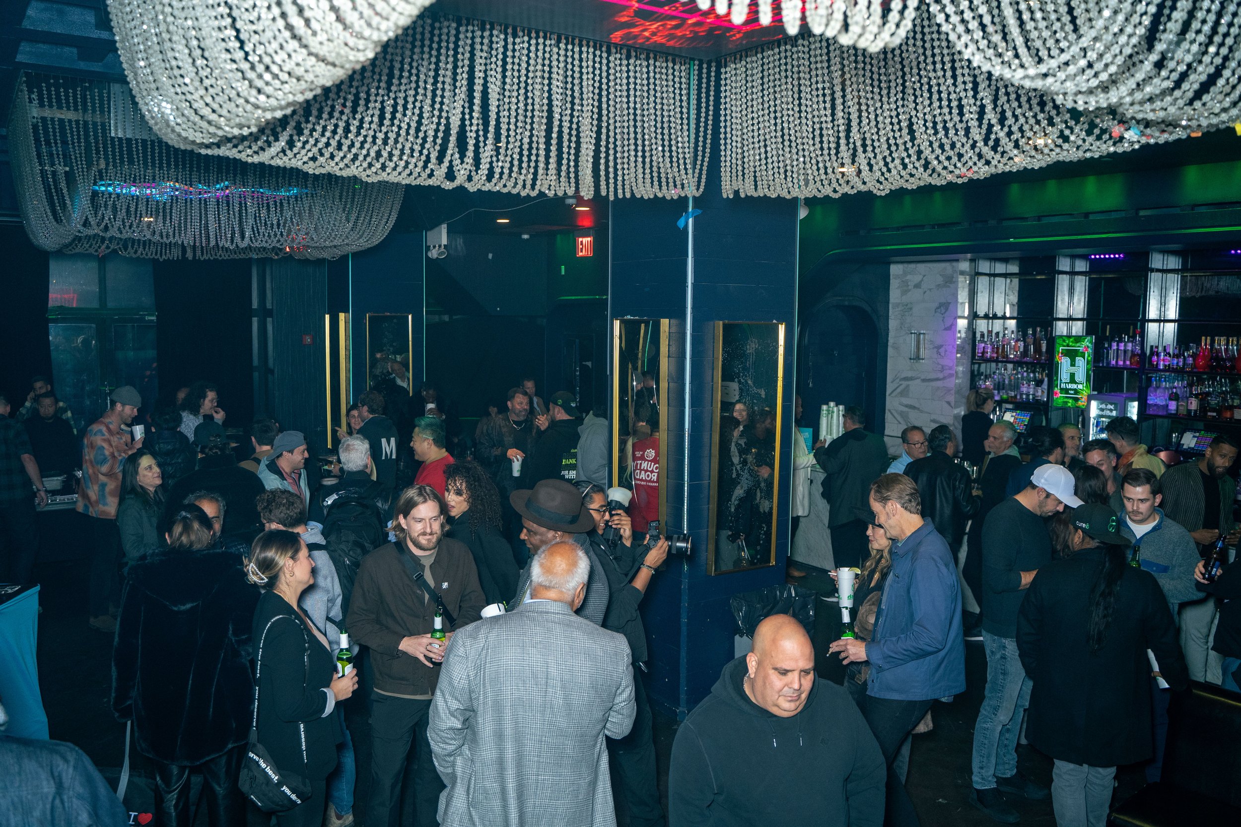 Crowd of people socializing inside a bar or nightclub, with a bar counter and colorful lighting in the background.