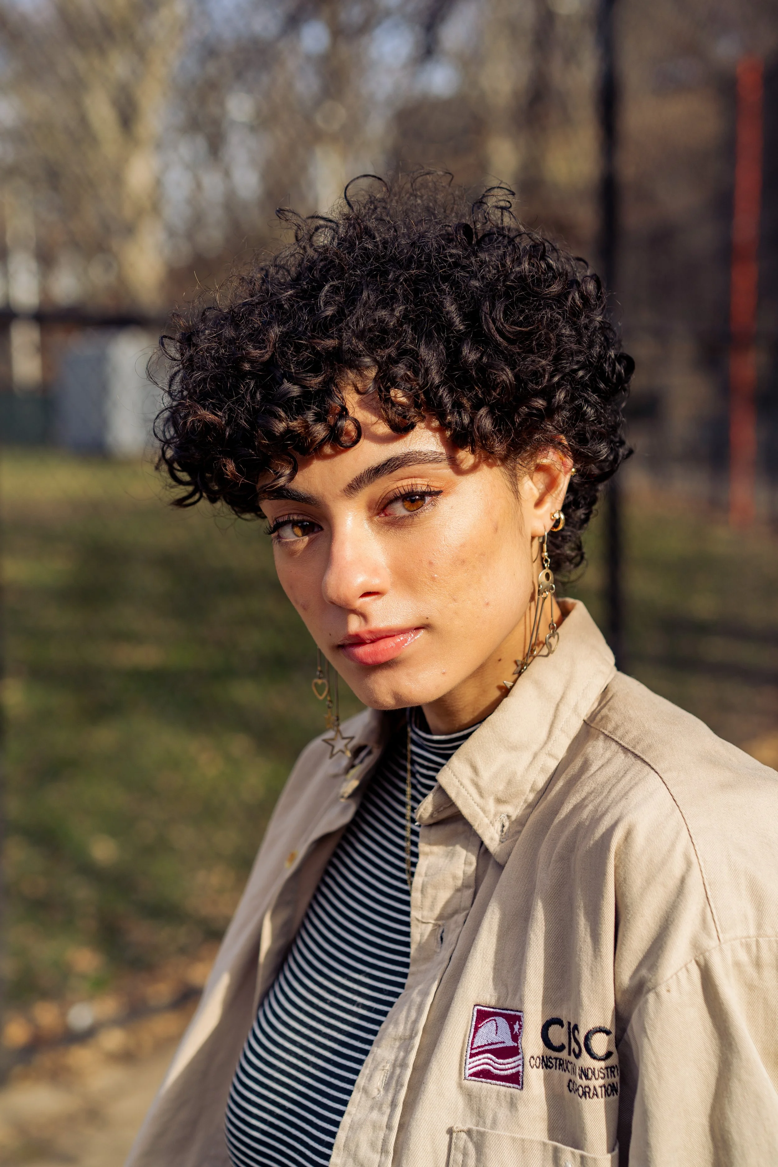 A young woman with curly short hair in Harlem and gold hoop earrings stands outdoors on a sunny day, wearing a beige work shirt with a logo and a black-and-white striped shirt underneath.
