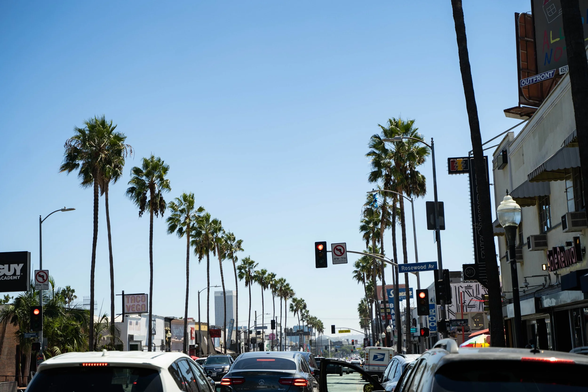 Street view with palm trees, cars, and storefronts on a sunny day in Los Angeles.