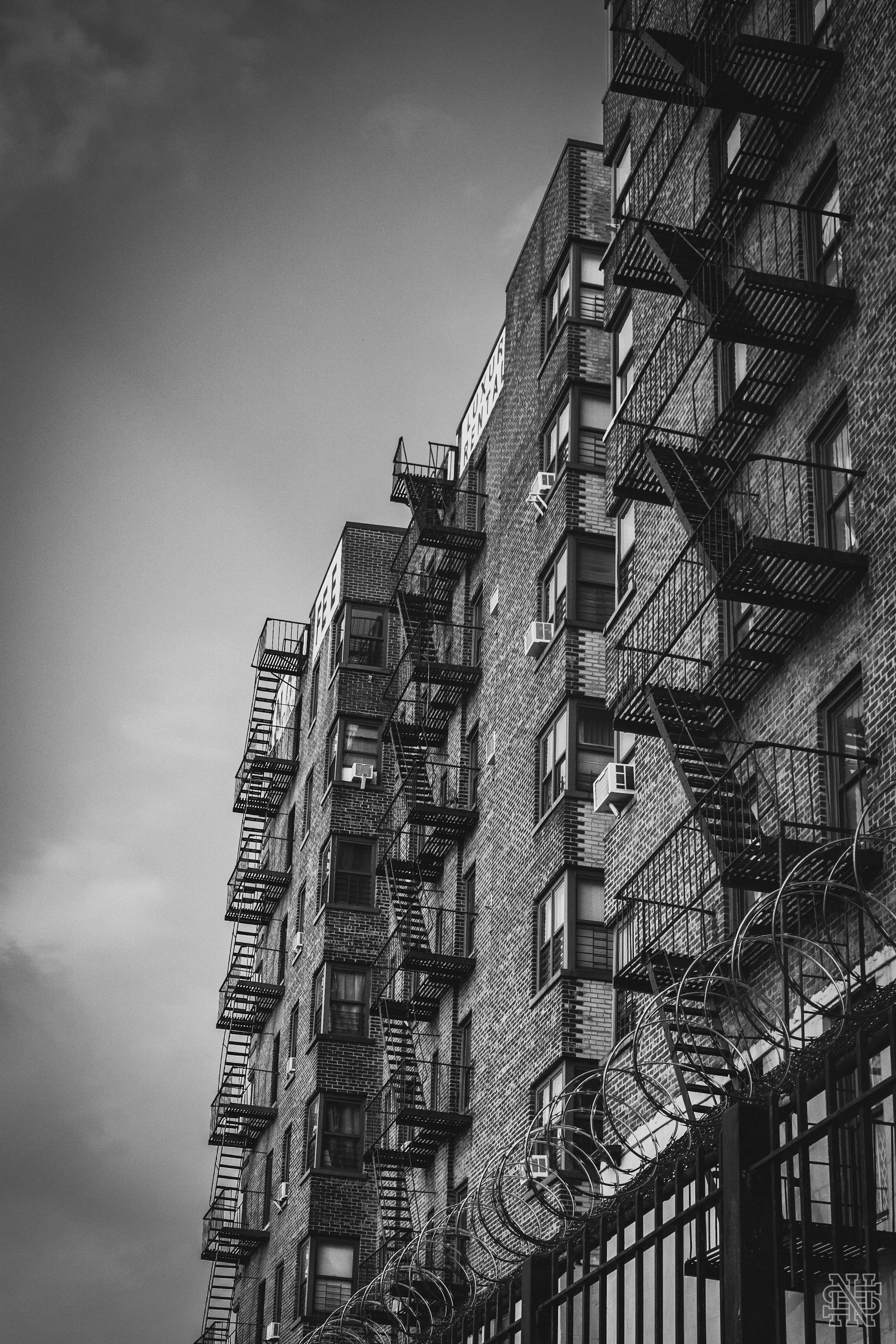 Black and white photo of a high-rise apartment building with fire escape stairs on the exterior and a barbed wire fence at the bottom.