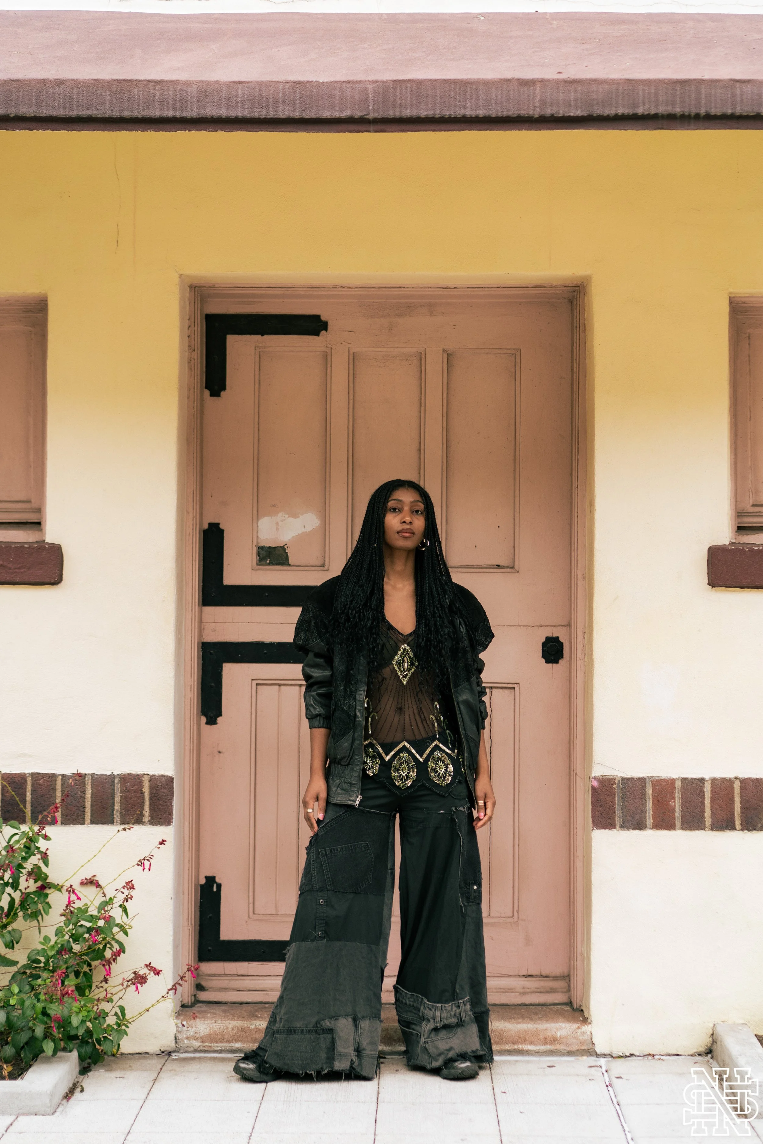 A woman with long braided hair wearing a black jacket, sheer top with embellishments, and oversized patchwork pants standing in front of a pink door and yellow wall.