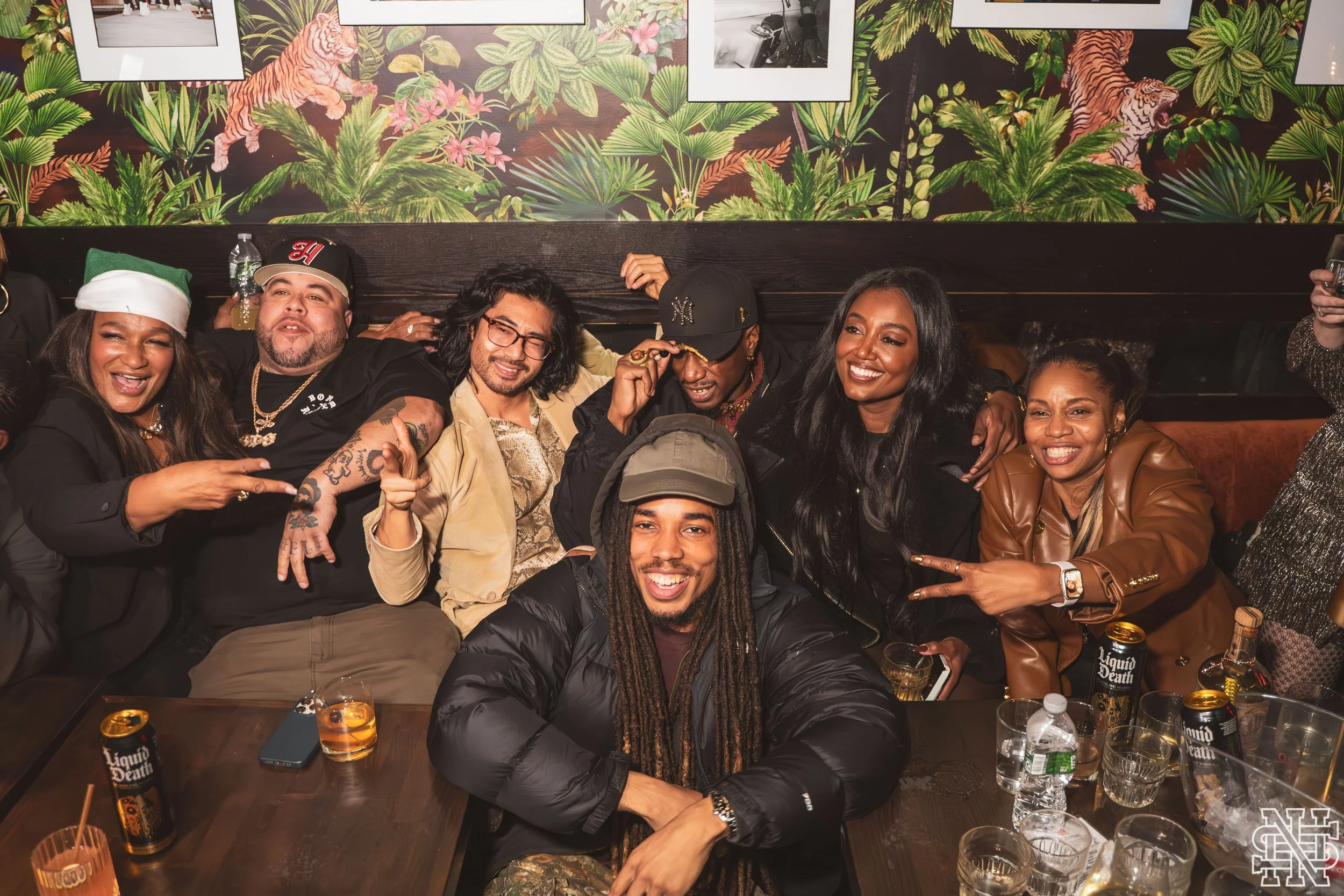 A group of nine diverse friends in a restaurant celebrating, with some making peace signs and smiling. The background features a jungle-themed mural with tigers and tropical plants. The table has drinks and cans of Liquid Death water.