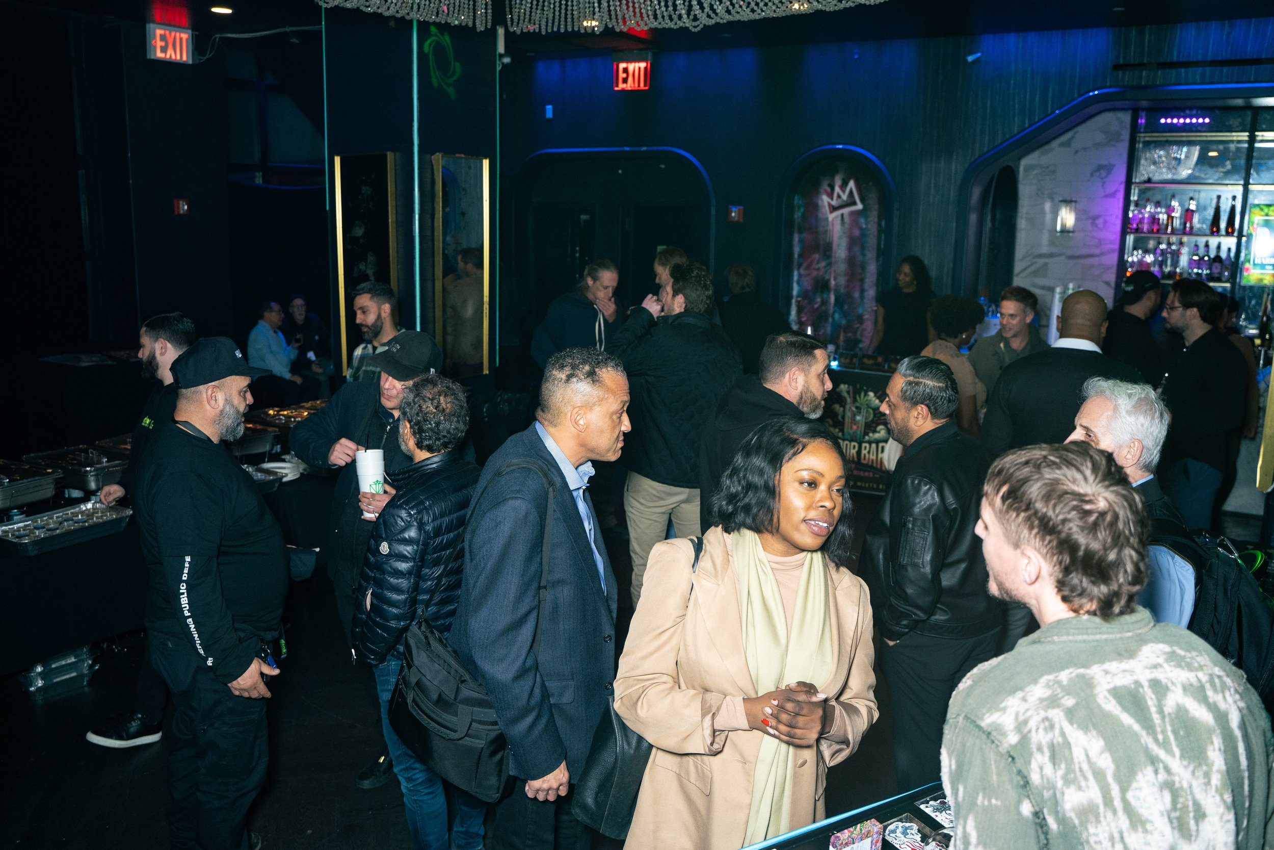 People gathering at a nightclub bar, engaging in conversations, with a DJ or bartender visible, colorful lighting, and Exit signs on the wall