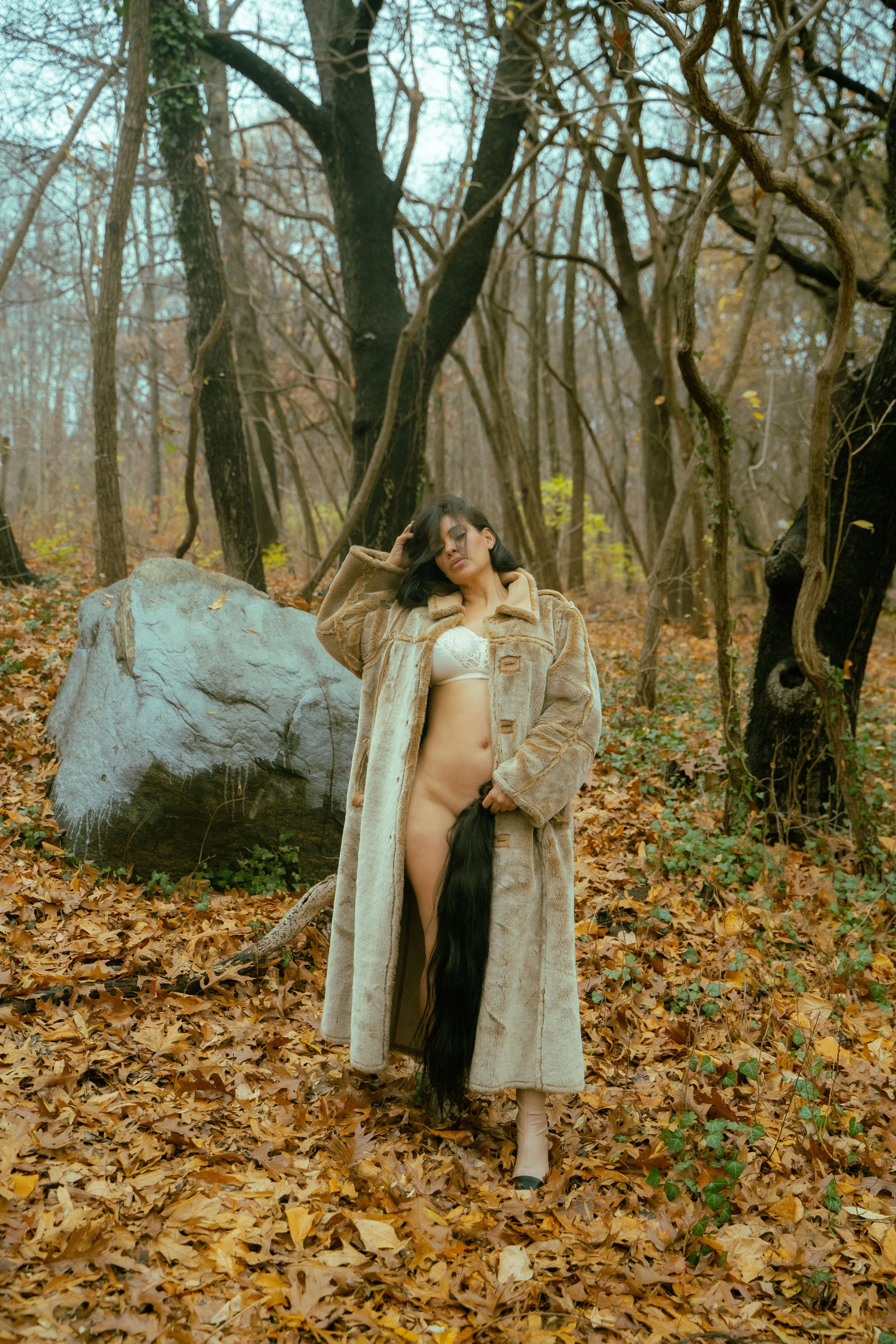 A woman standing in a forest covered with fallen autumn leaves, wearing a long beige coat, lingerie, and high heels, with long dark hair and a large rock beside her.