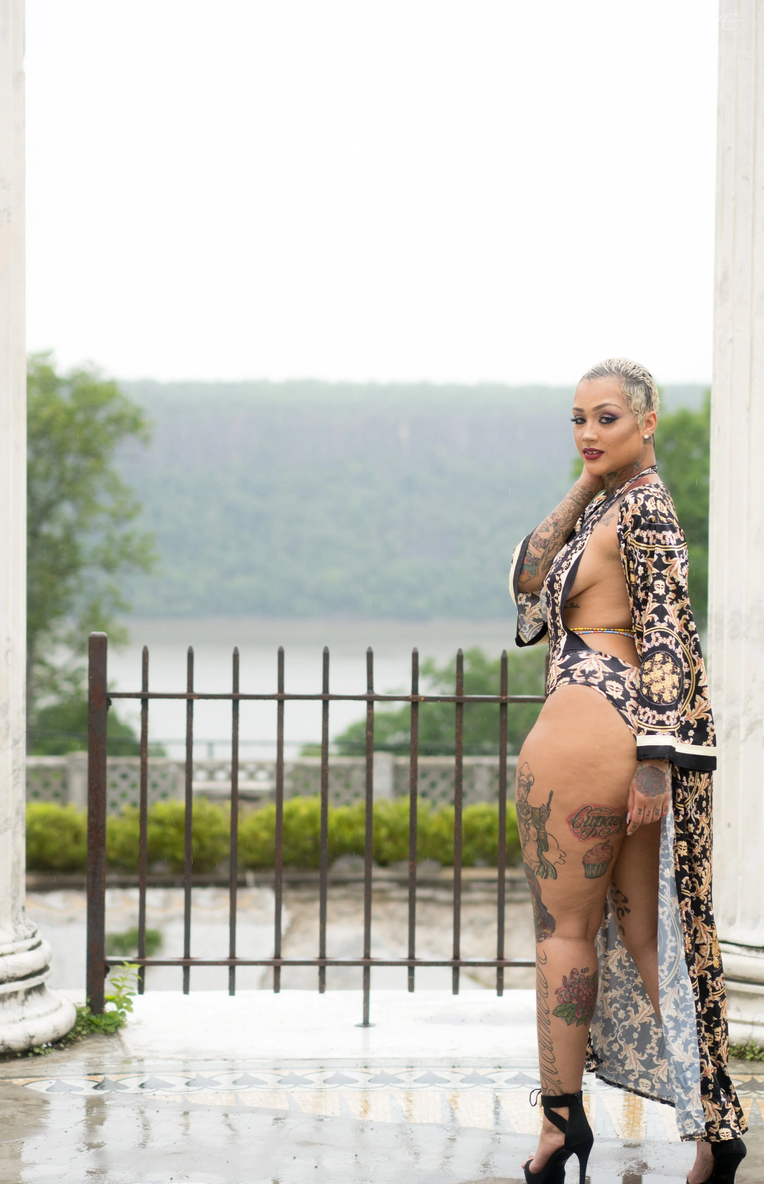 A woman with tattoos and short blonde hair standing on a balcony with columns, overlooking a body of water and green landscape, wearing a revealing dress and high heels.