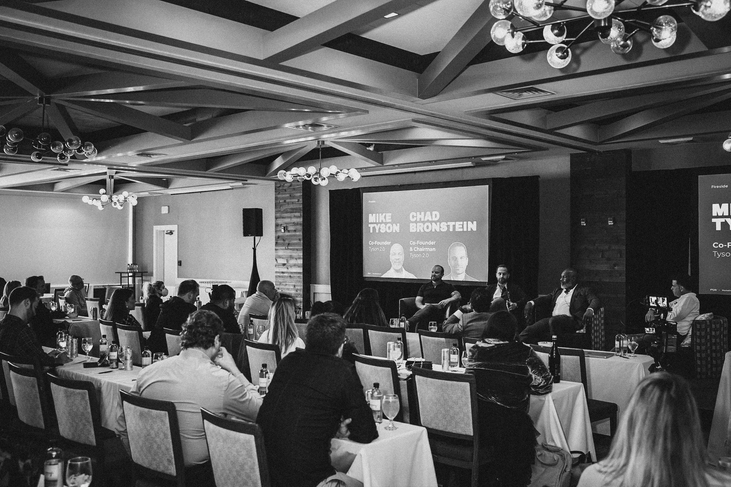 A black and white photo of a panel discussion in a conference room with an audience seated at tables. Four men are seated on a stage, engaged in discussion, with a large screen behind them displaying their names: Mike Tyson and Chad Bronstein.