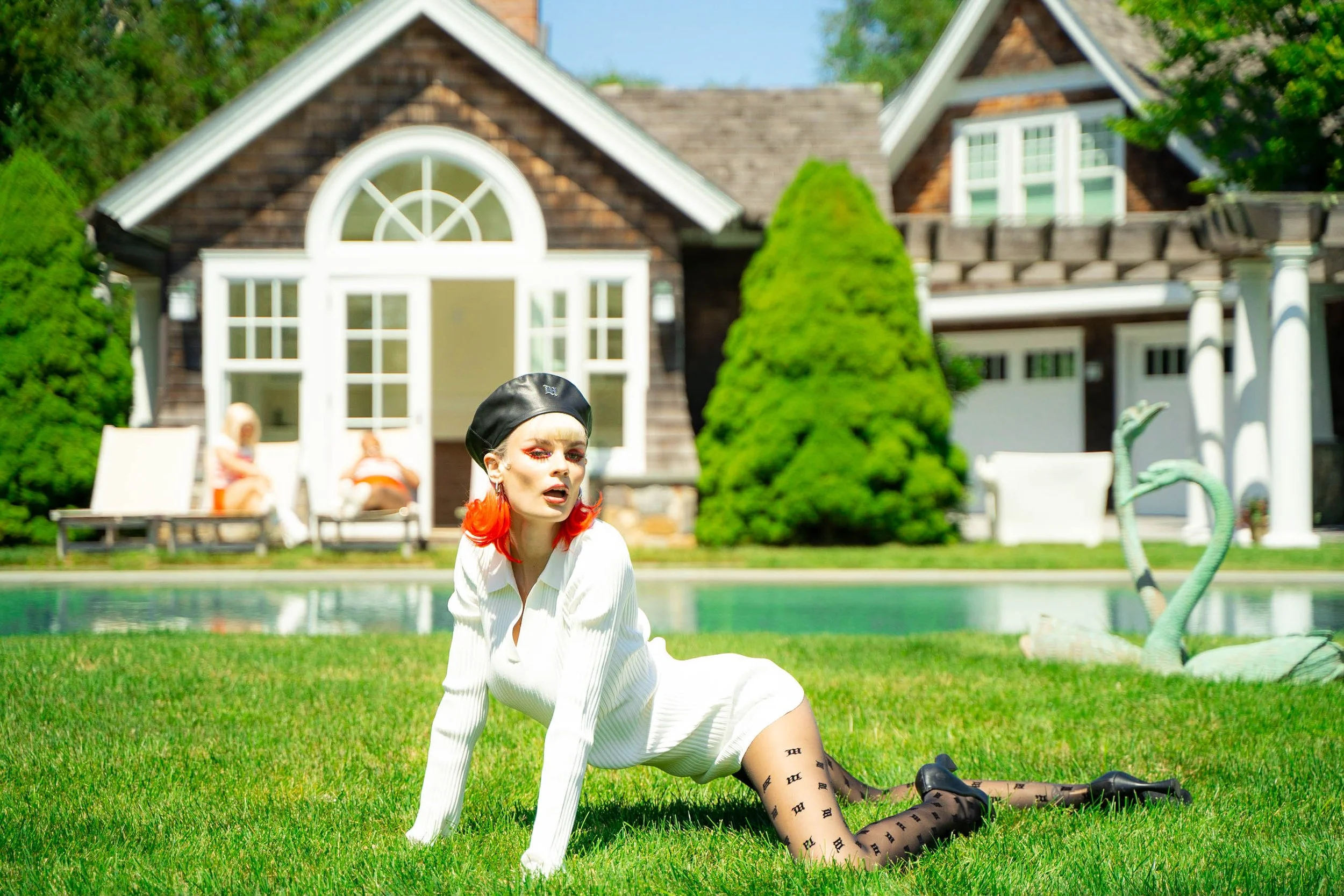 A woman with red hair and red earrings, dressed in a white outfit with black patterned tights and black shoes, kneeling on green grass in front of a house, with children and outdoor furniture in the background and a swimming pool with a decorative sw