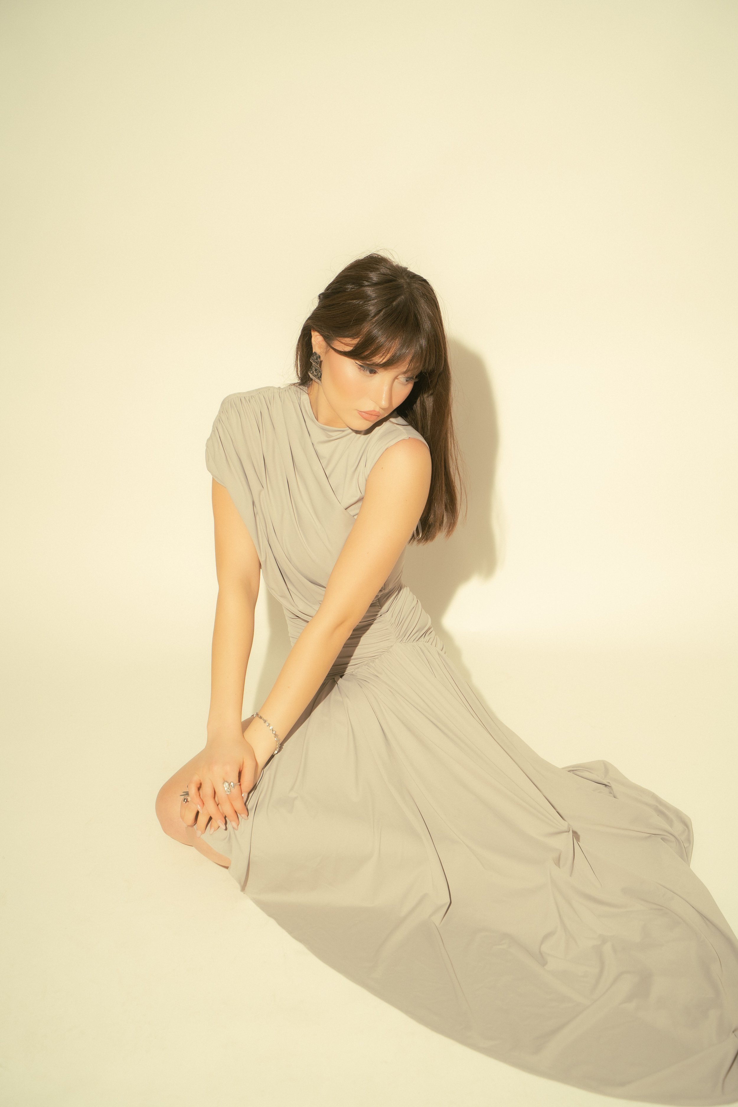 A woman with dark hair in a beige dress sitting on the floor against a plain light-colored background.