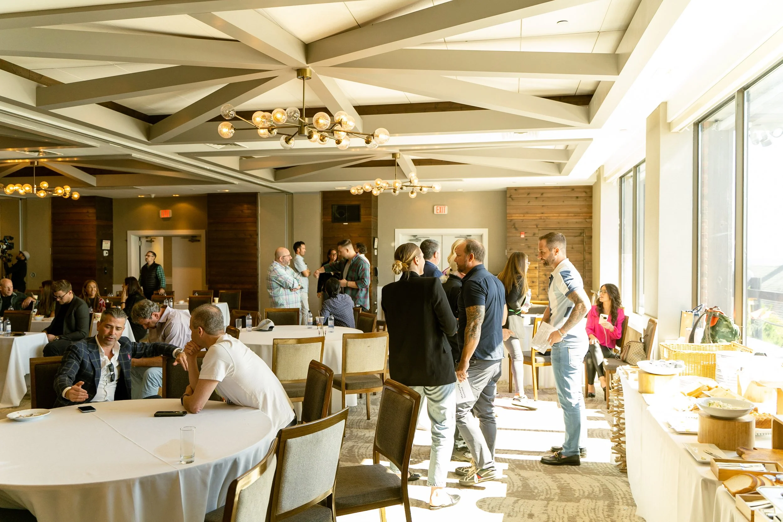 People gathered in a well-lit room with large windows, some standing and talking, others seated at round tables, with a buffet table on the right side. The ceiling has modern light fixtures.