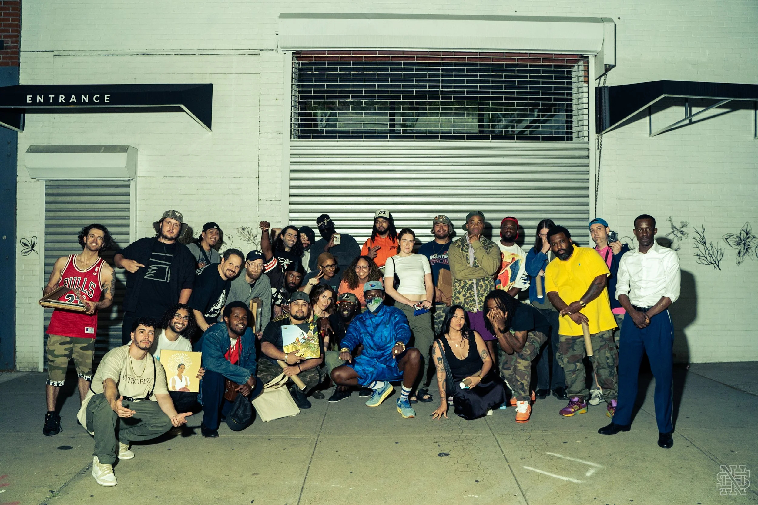 A large group of diverse people posing in front of a closed garage door with a white brick wall, some holding items, some making gestures, some kneeling or standing, in an urban setting at night.