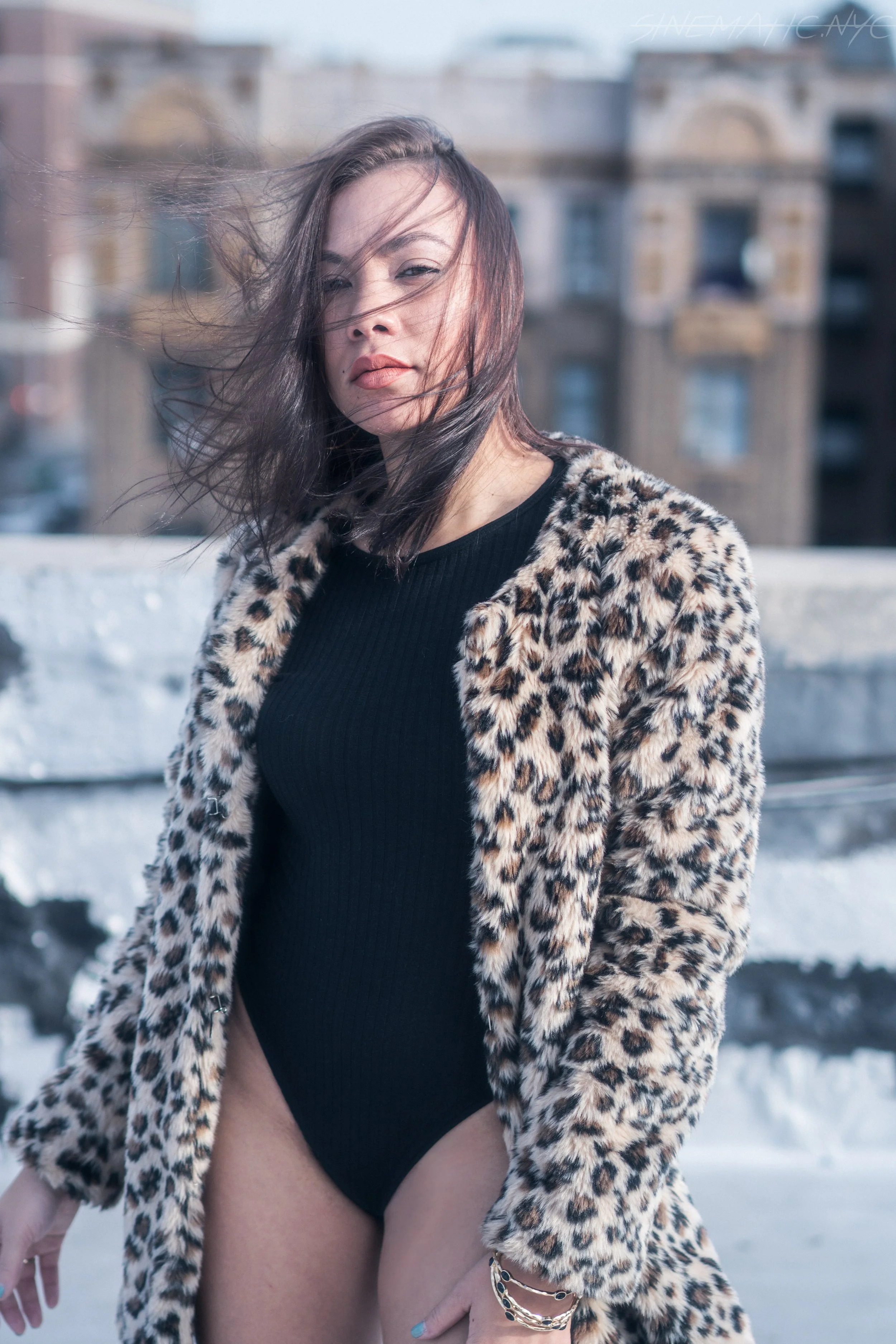A woman with brown hair outside, wearing a leopard print coat over a black top, with wind blowing her hair.