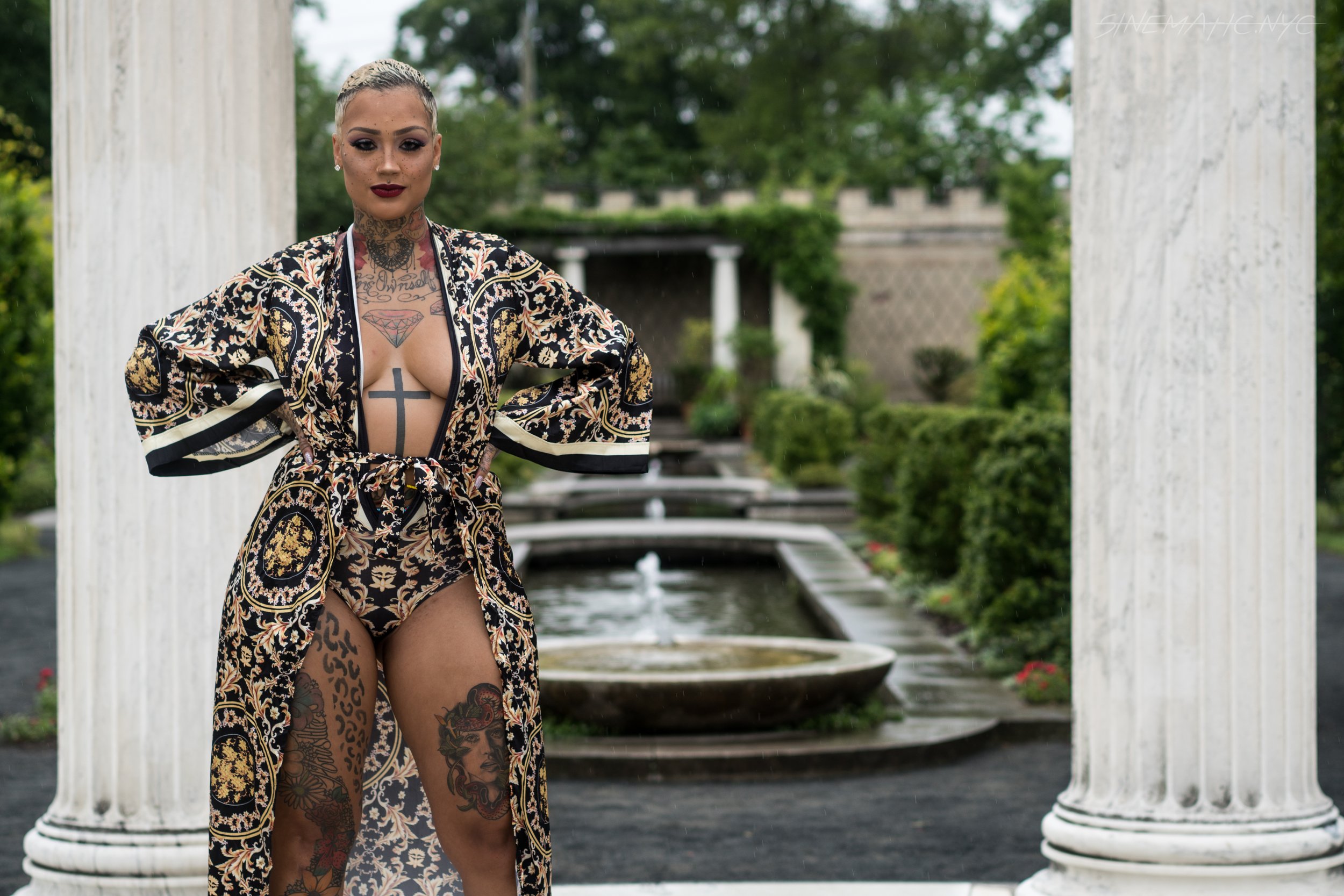 A woman with multiple tattoos, wearing a black and gold patterned robe open to reveal tattoos on her chest and thighs, standing outdoors near white classical columns with a fountain and lush greenery in the background.