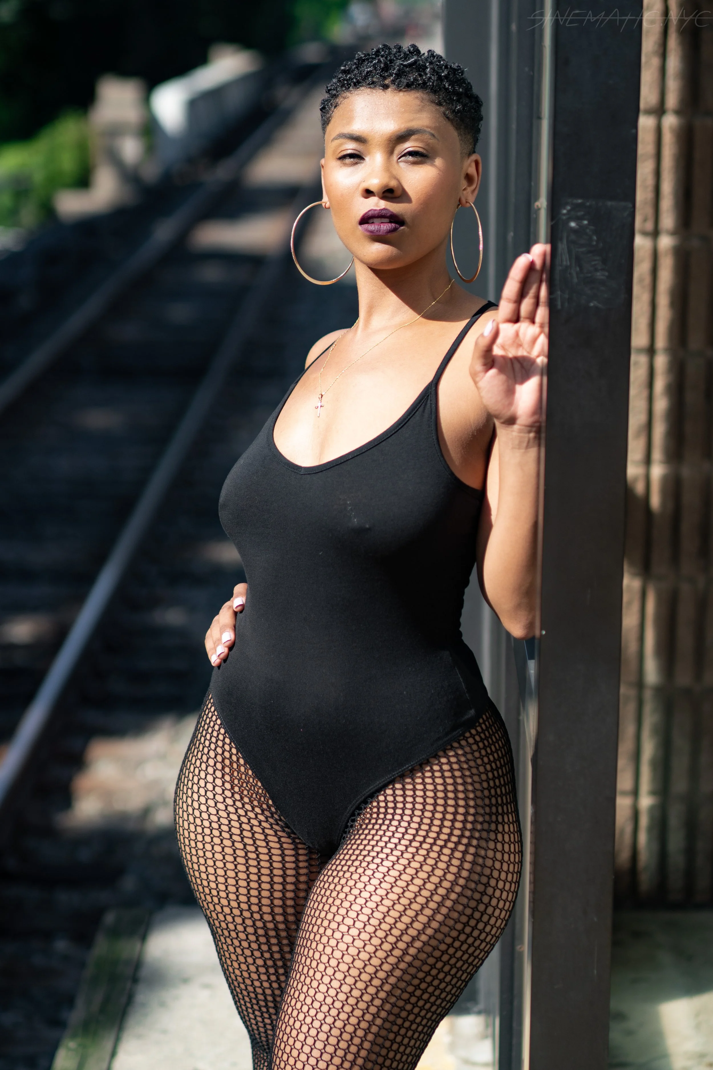A woman standing outdoors next to a metal pole, wearing a black tank top, fishnet stockings, large hoop earrings, and a necklace with a cross pendant. She is making eye contact with the camera and holding her hand near the pole.