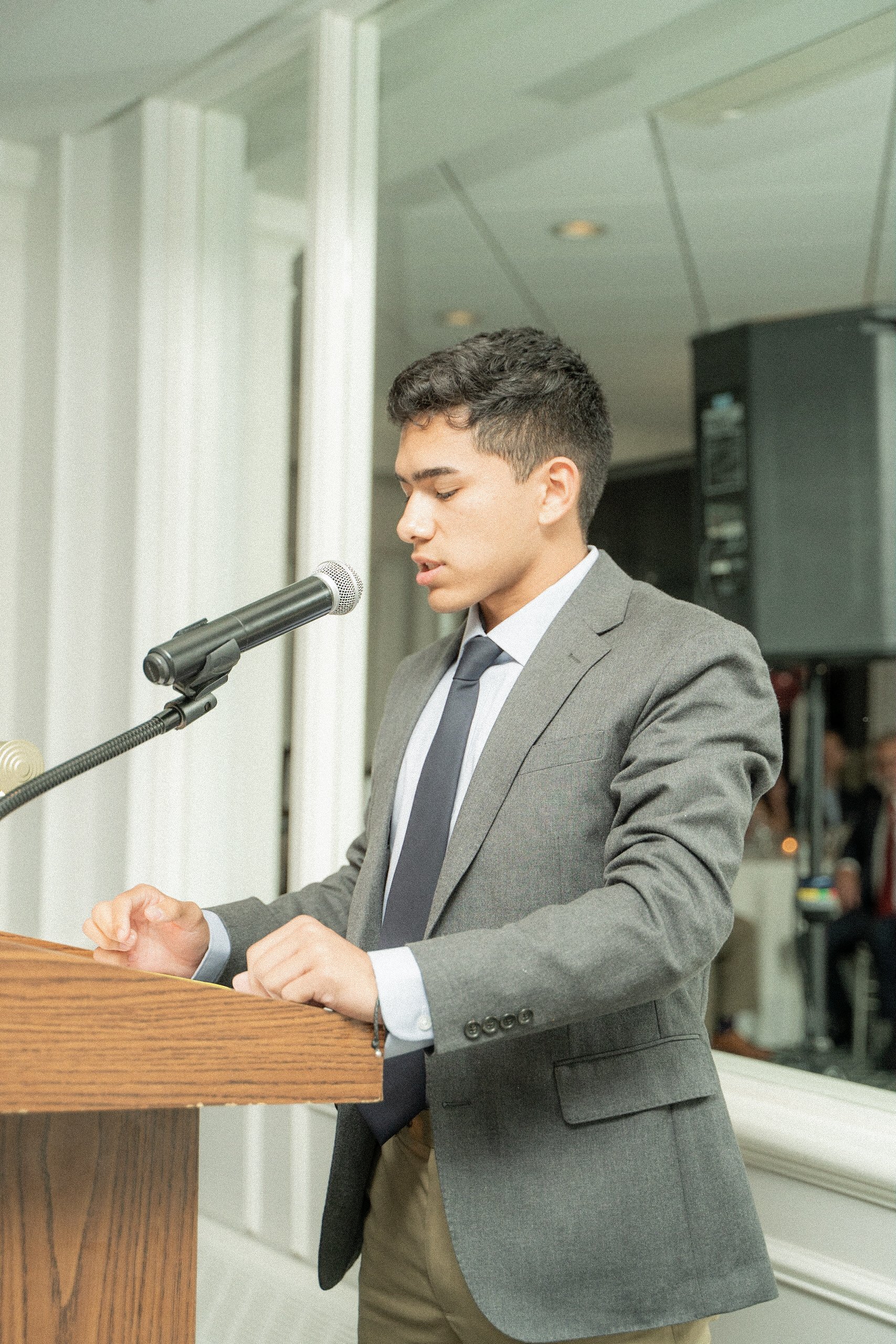 A young man in a gray suit and tie, standing at a wooden podium, speaking into a microphone at an indoor event.
