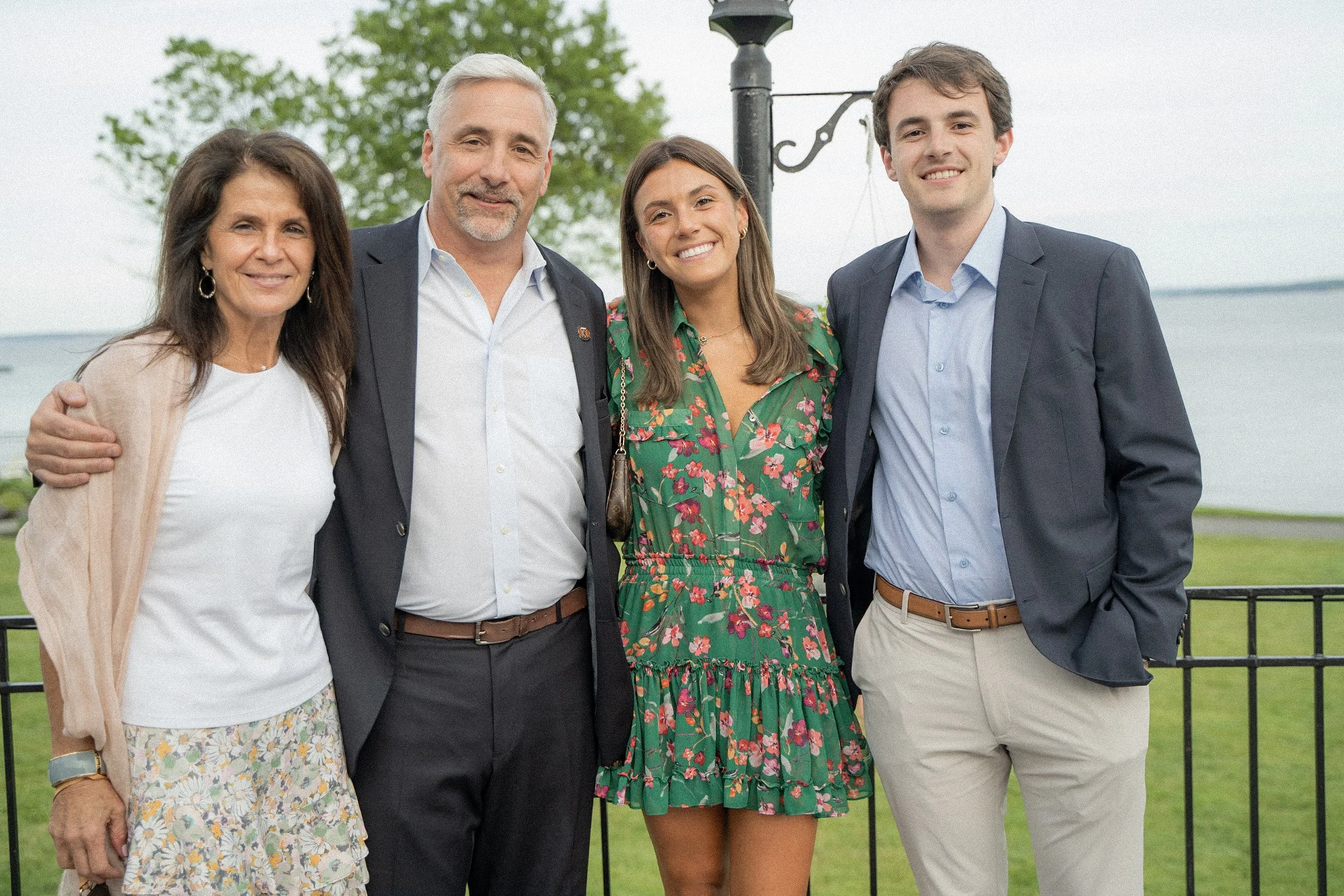 Four people standing outdoors near a body of water, smiling and posing for a photo. Two women and two men, dressed in casual and semi-formal clothing, with trees and a railing in the background.