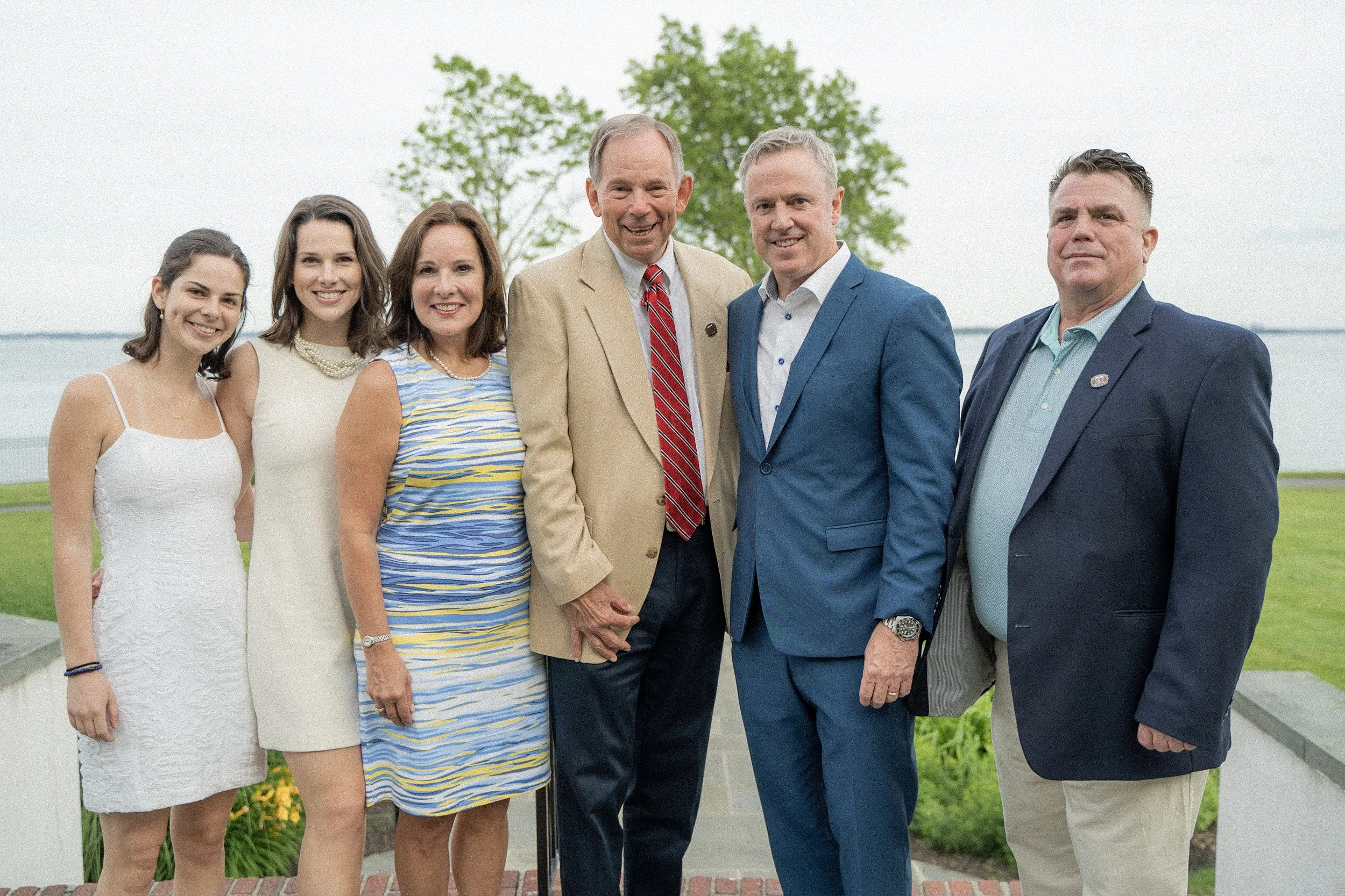 Group of seven people standing outdoors near water with green trees in the background, dressed in formal and semi-formal attire.