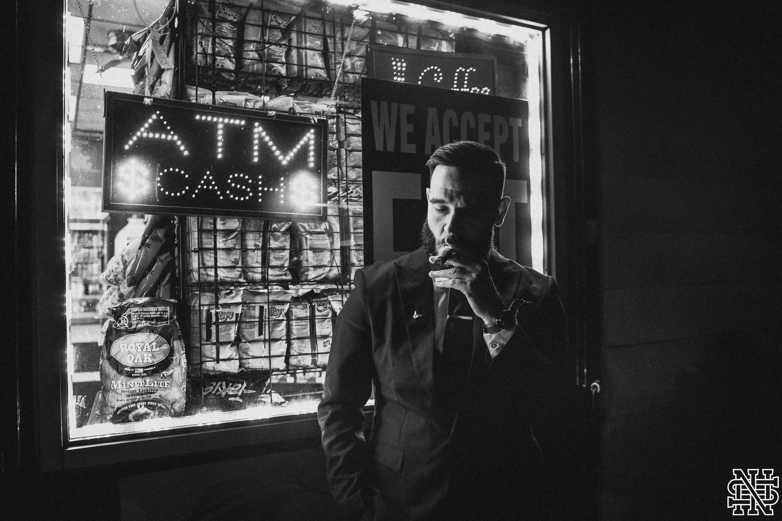 A man with a beard and mustache in a suit smokes a cigar outside a convenience store at night. Behind him, a lit sign reads "ATM CASH" and a larger sign says "WE ACCEPT." The store window displays snack bags and a poster with the number 716. The scen