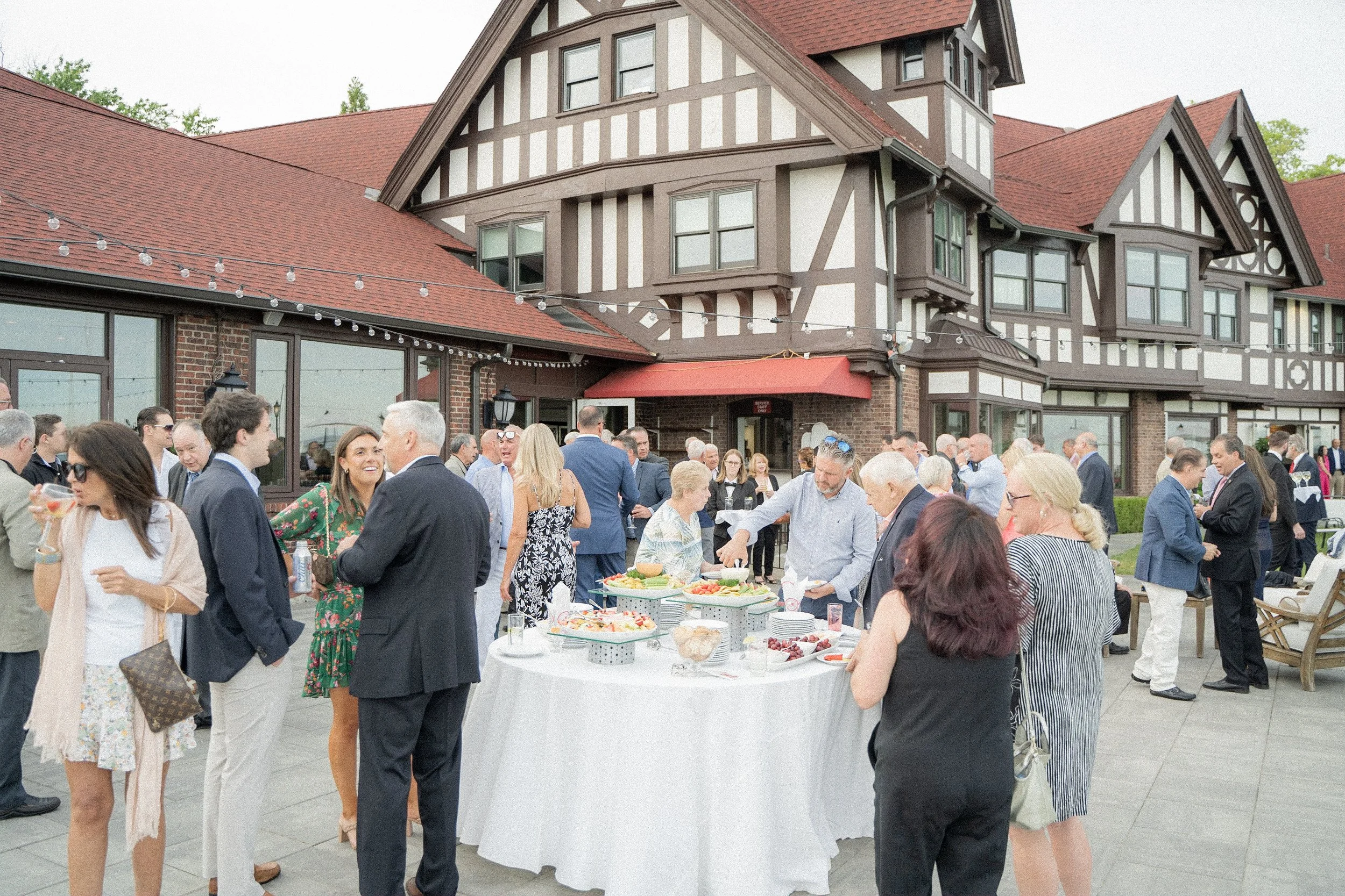 People socializing at an outdoor gathering on a patio in front of a large, traditional-style building with red roof and timber framing.