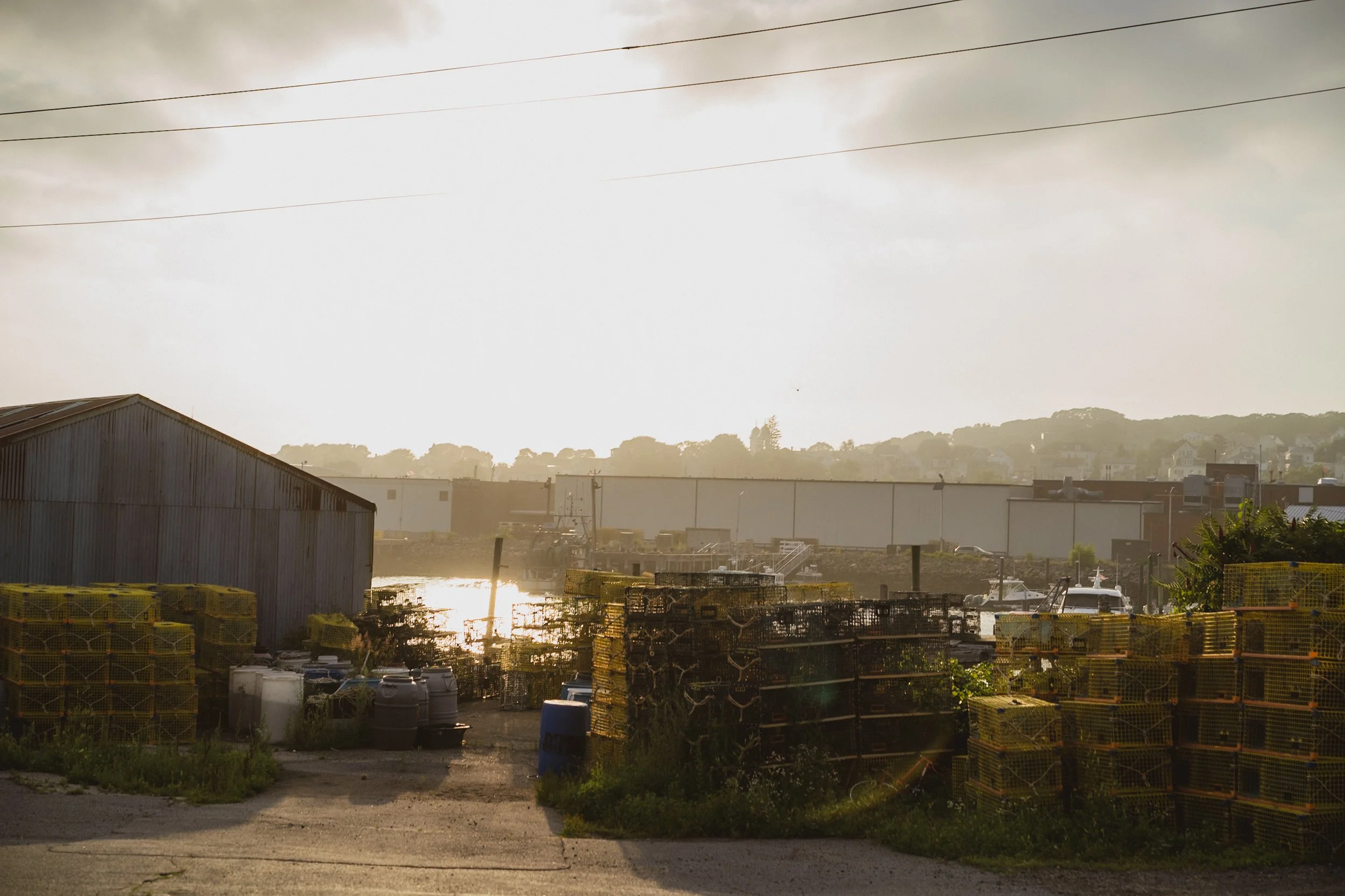 An industrial area with stacks of lobster traps, a shed, and a boat docked by the water, during sunset with a cloudy sky.