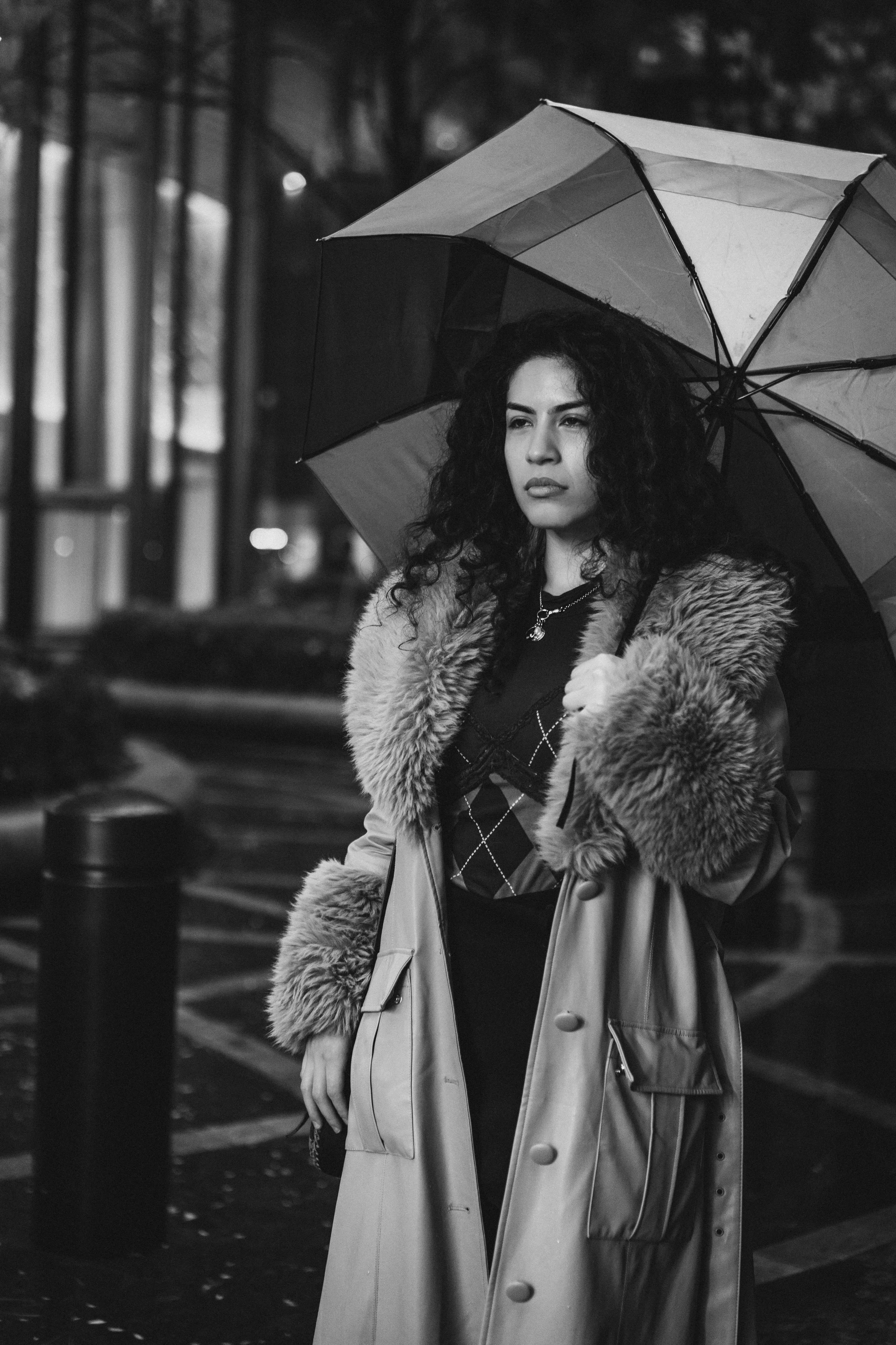 A woman stands outdoors at night holding an umbrella, wearing a long coat with faux fur trim on the collar and cuffs, with a serious expression on her face.