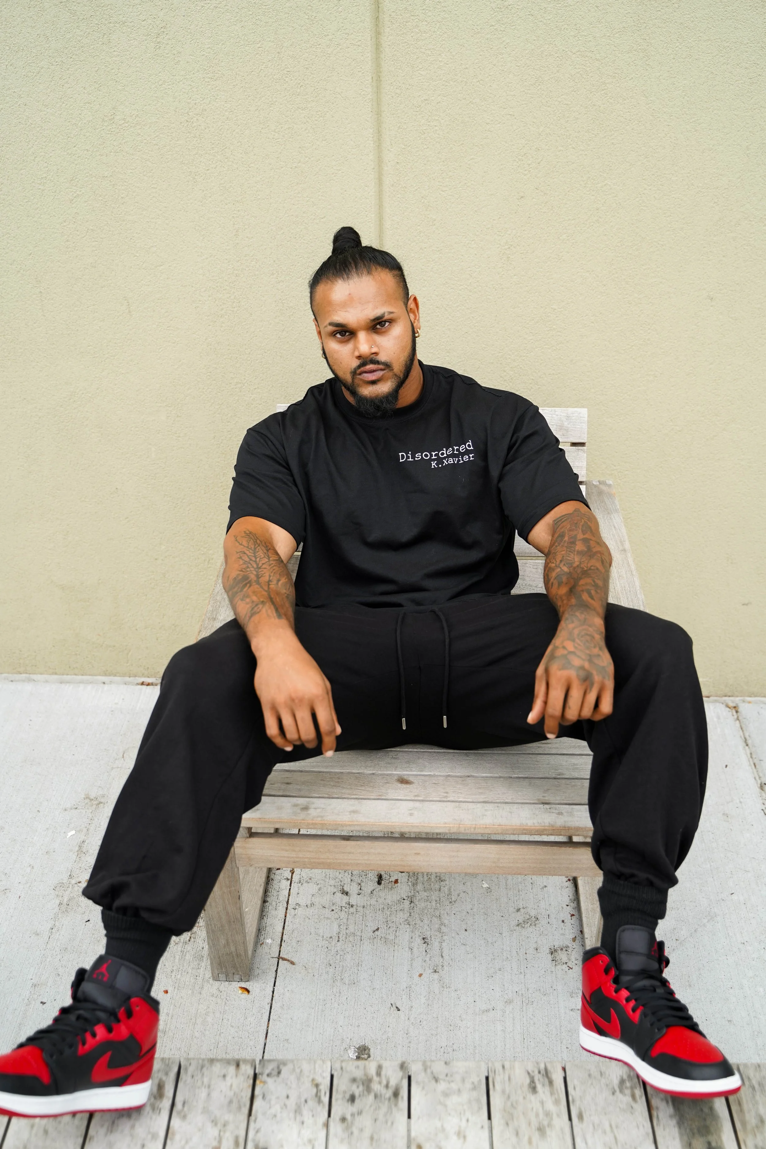 A young man with dark skin, tattoos, and a beard, sitting on a wooden bench against a plain beige wall. He is wearing a black t-shirt with text, black pants, and red and black sneakers, with a serious expression on his face.