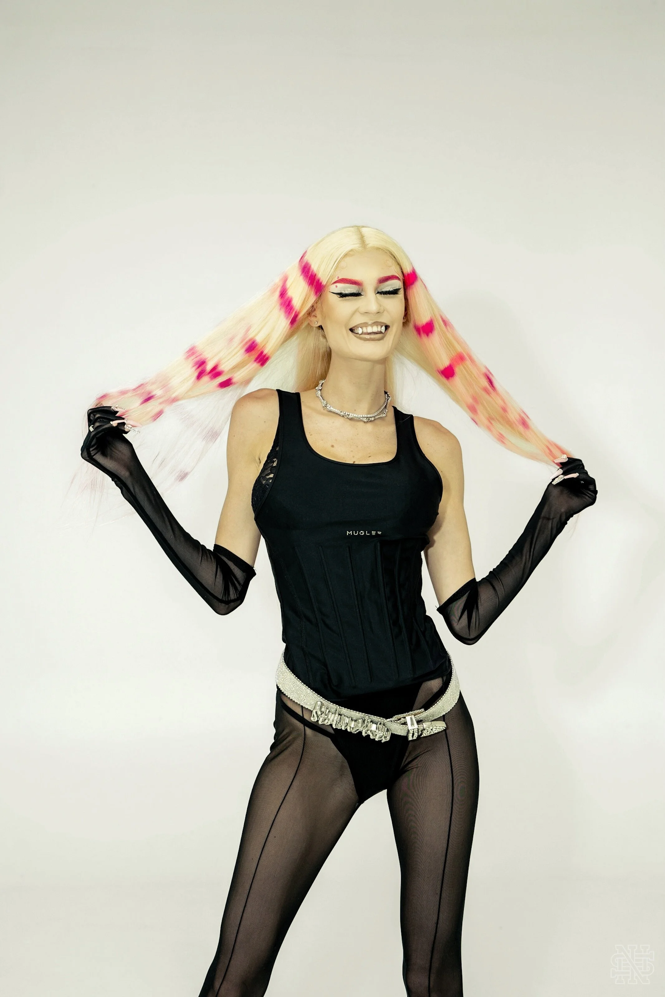 Person with long blonde hair with pink streaks, wearing a black sleeveless top, black sheer tights, and long black gloves, smiling and holding her hair, standing against a plain white background.