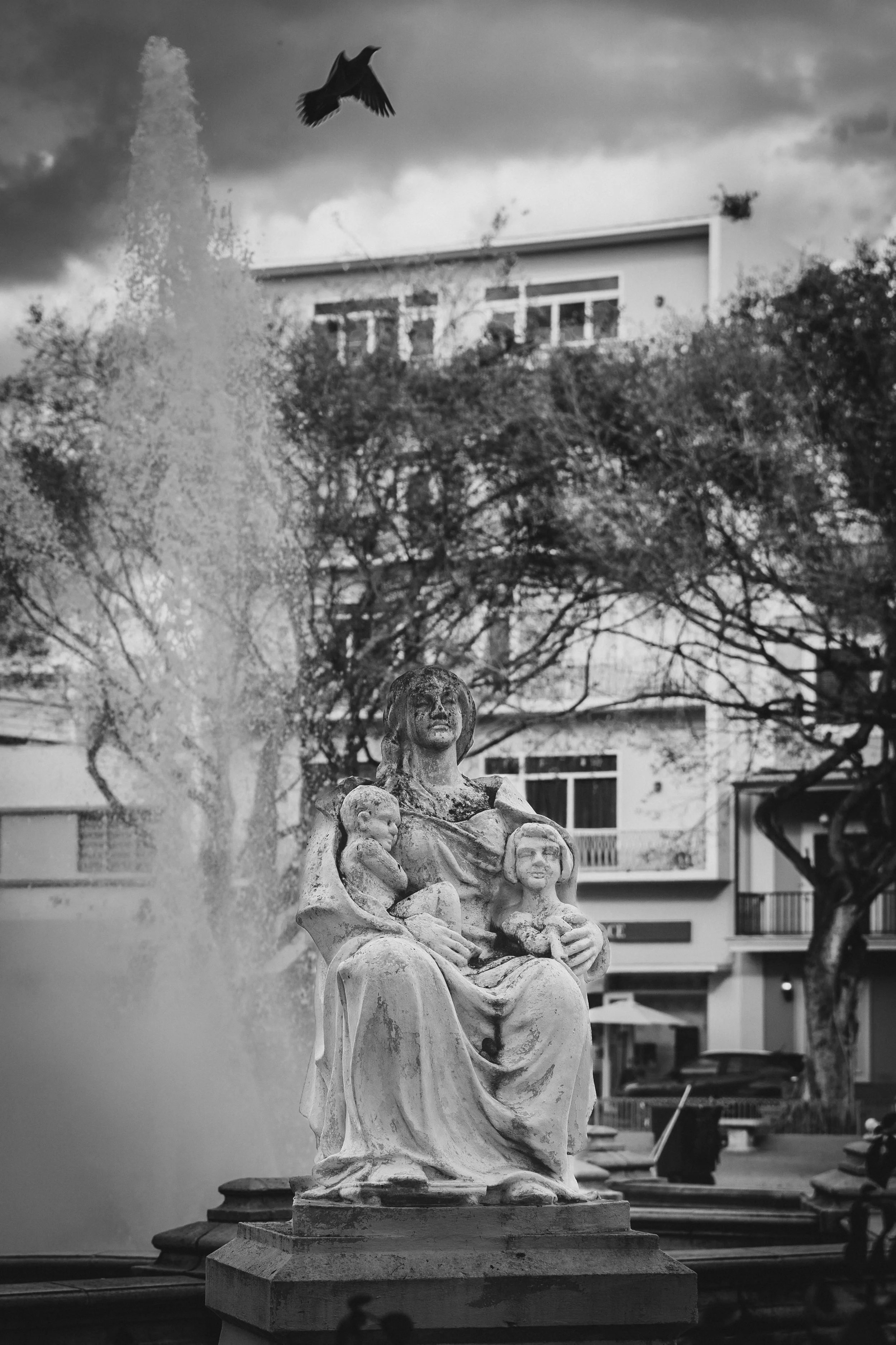 A black-and-white photo of a fountain with a statue of a woman holding two children, with a fountain spray on the left and a bird flying above. There are trees and buildings in the background.