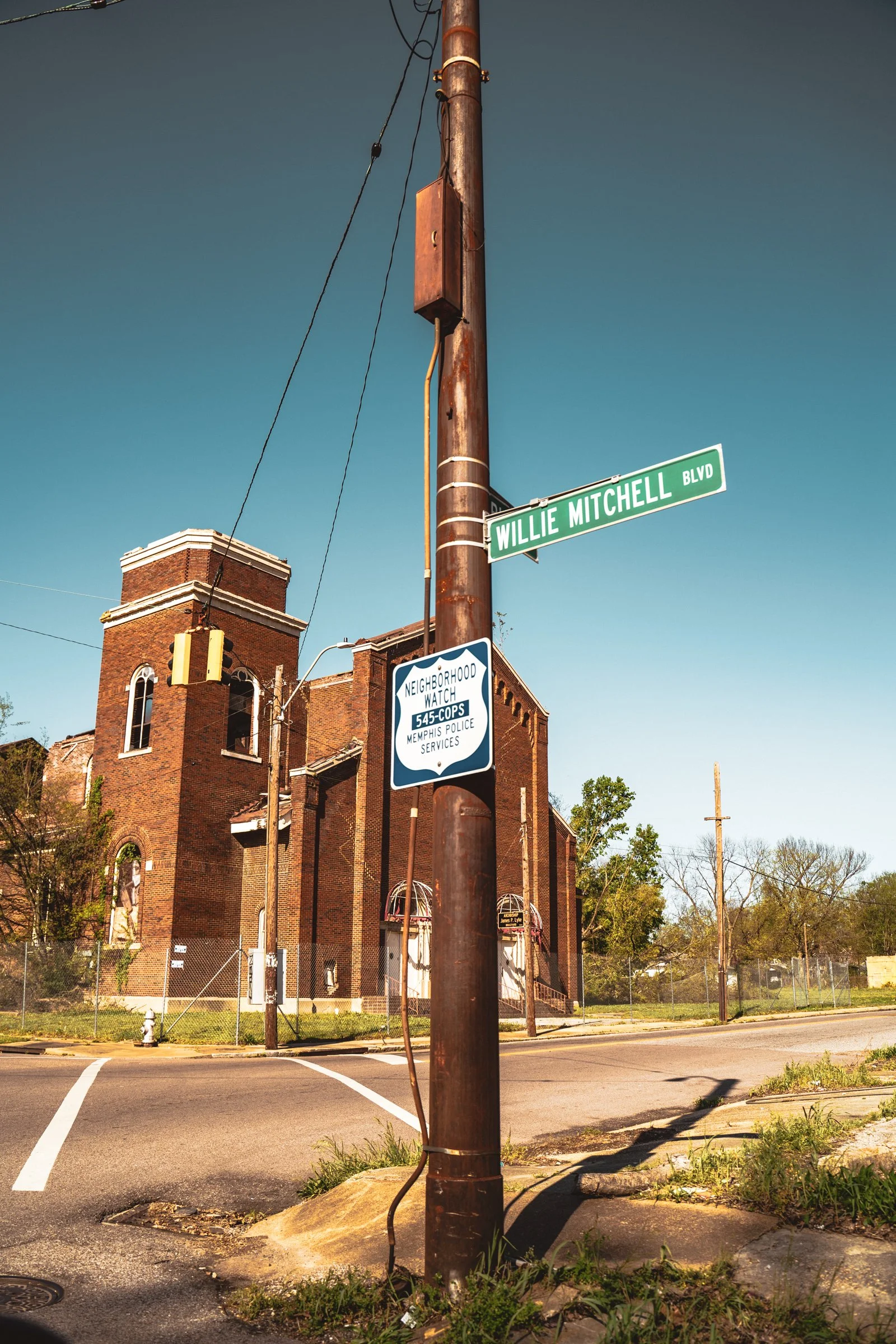 Street corner with a street sign for Willie Mitchell Boulevard, a utility pole with a neighborhood watch sign, and a brick building with a rounded tower in the background. Clear blue sky and some trees are visible.