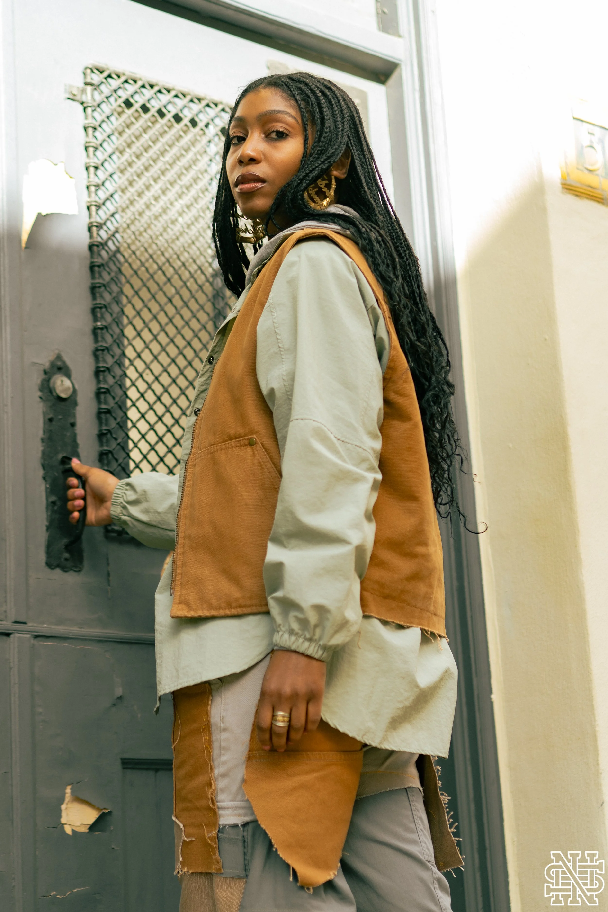 A young woman with long braided hair, wearing a beige and brown layered jacket, standing by a gray door with a grated window, holding the door handle.