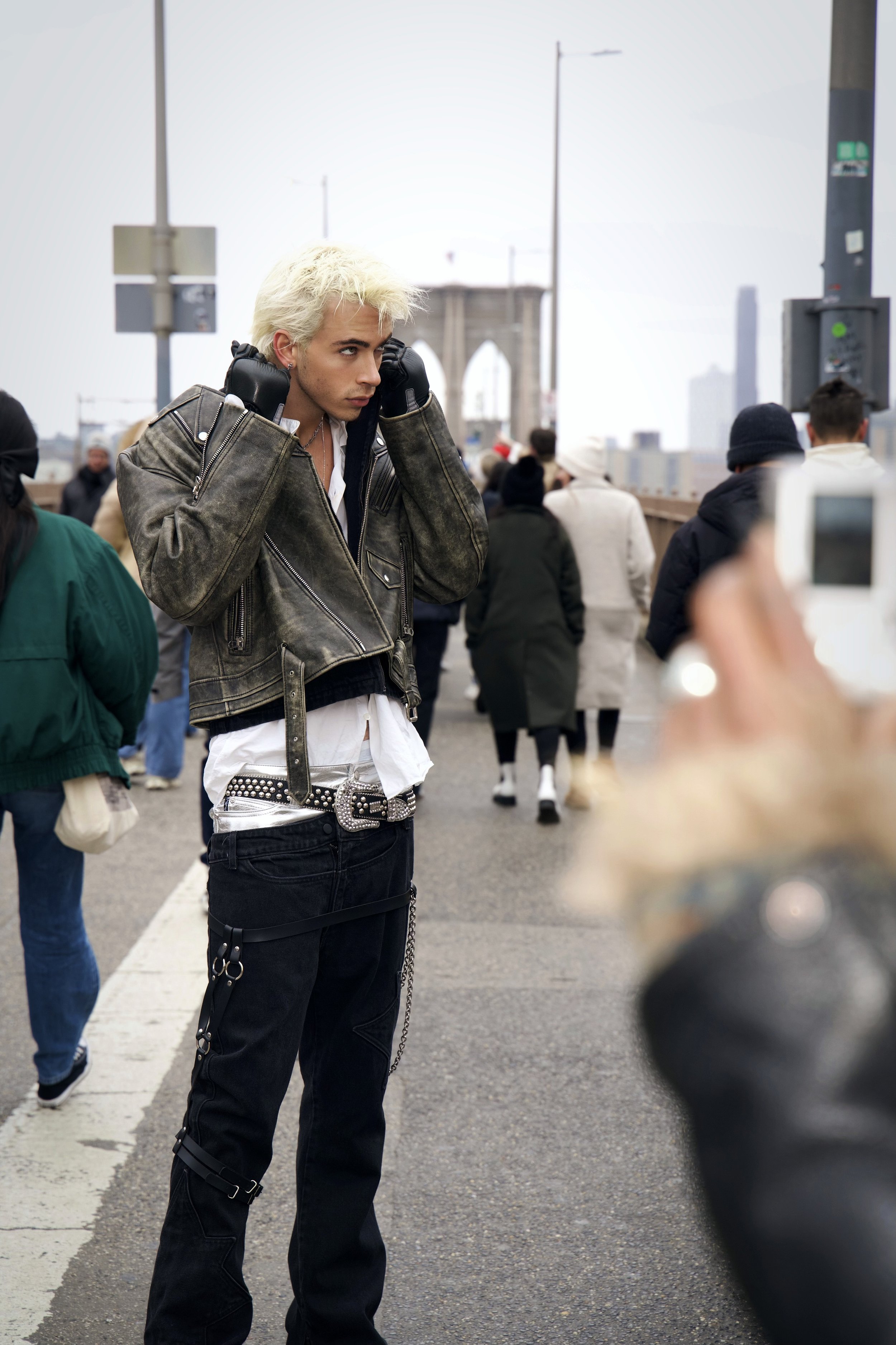 Young man with platinum blonde hair, wearing a leather jacket, white shirt, and black pants with chains and a studded belt, standing on a bridge in New York City, surrounded by pedestrians.