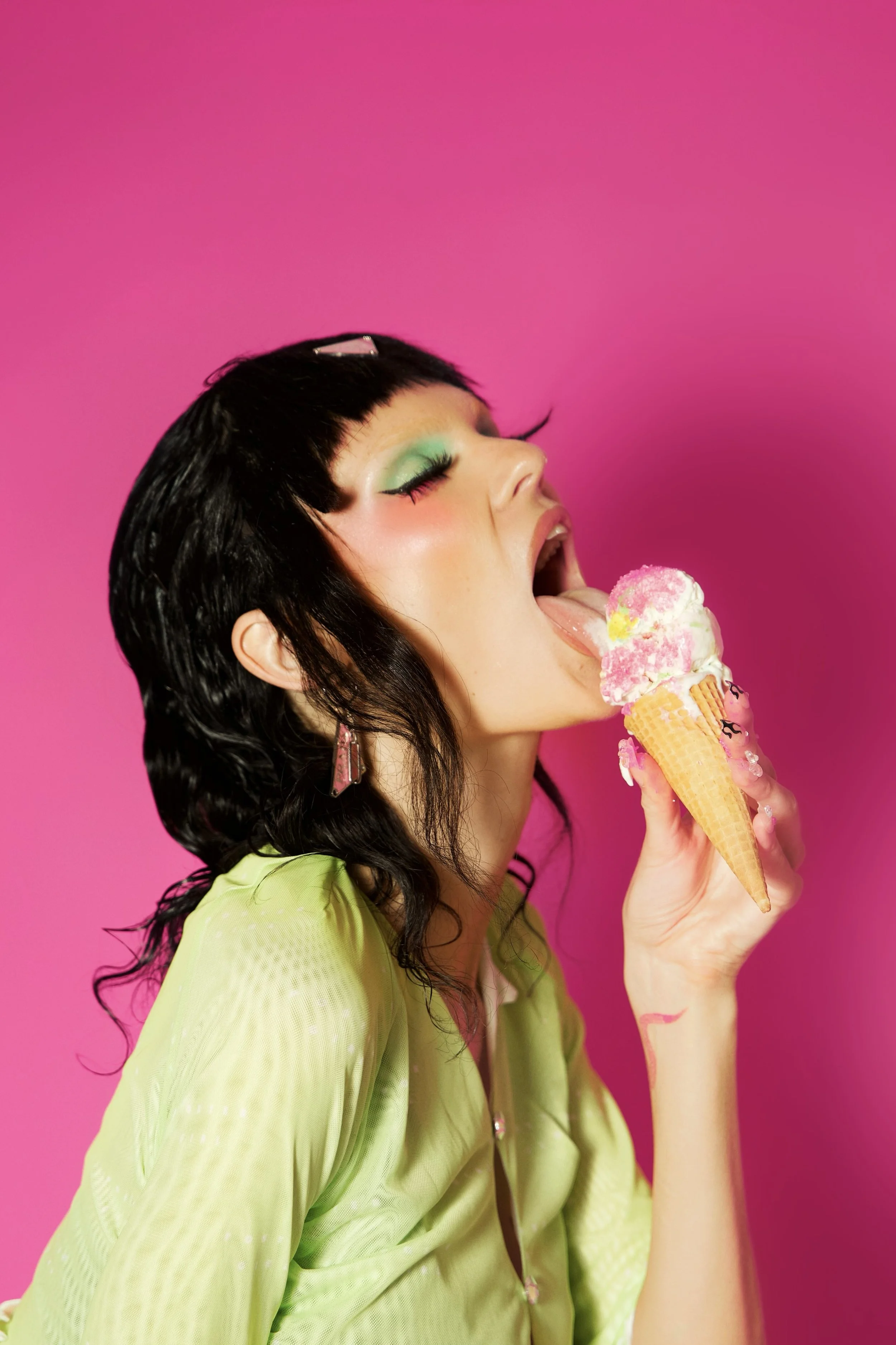 A person with dark hair and colorful makeup is licking a cone with pink and white ice cream against a pink background.